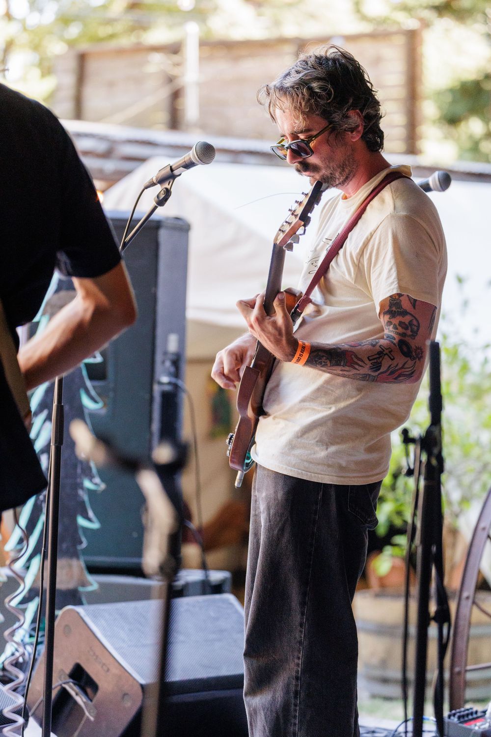 Tattooed guitarist performing on outdoor stage, wearing white t-shirt and sunglasses, playing electric guitar at music festival.