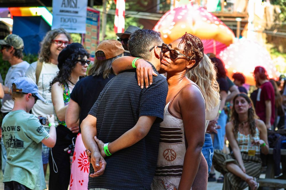 Couple kissing at outdoor festival surrounded by crowd of people in colorful summer attire.