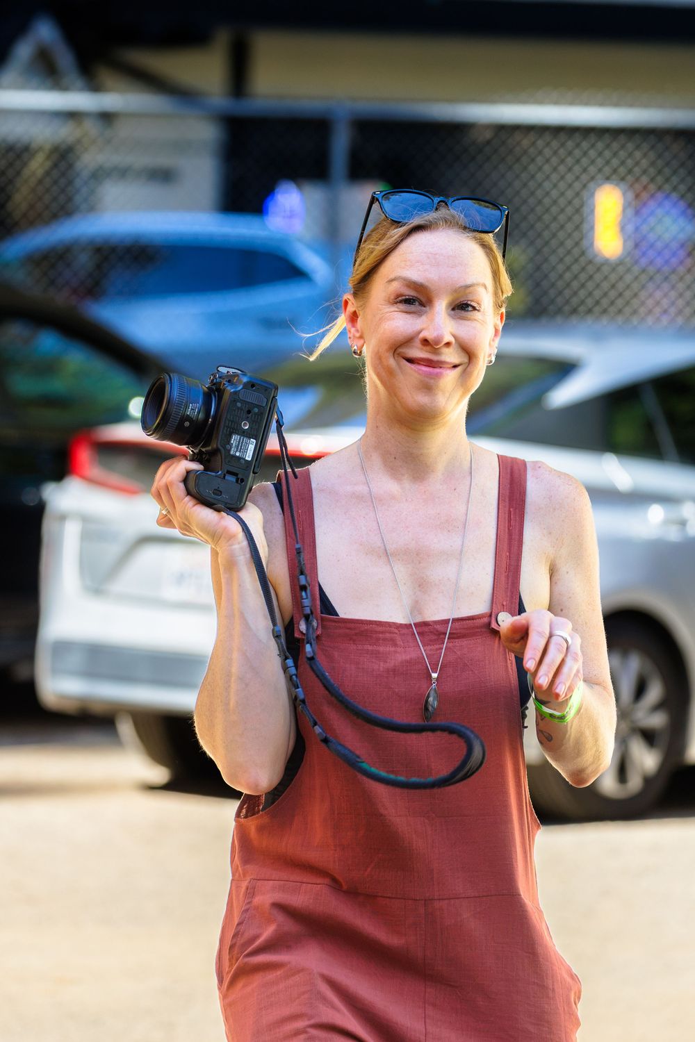 Smiling woman in red dress holding a camera outdoors near parked cars with sunglasses on her head.