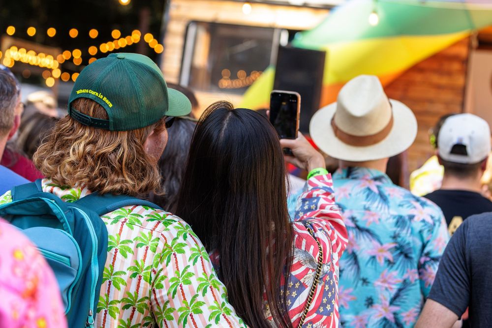 Crowd at outdoor festival wearing tropical shirts and hats, person taking photo with smartphone near colorful food truck.