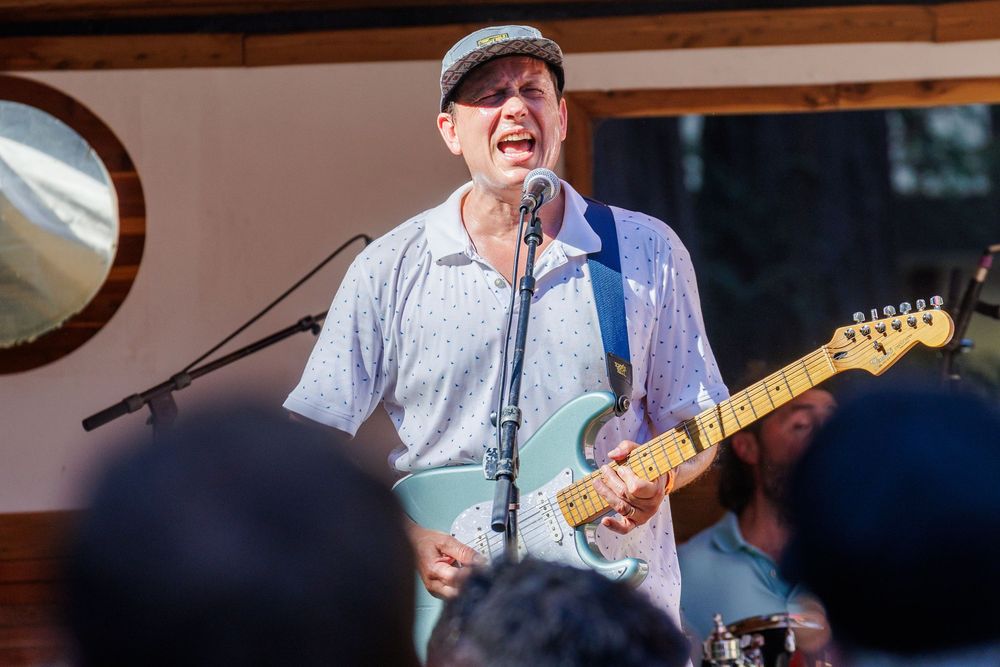 Male guitarist singing passionately on outdoor stage, playing mint green Fender Stratocaster at live music festival.
