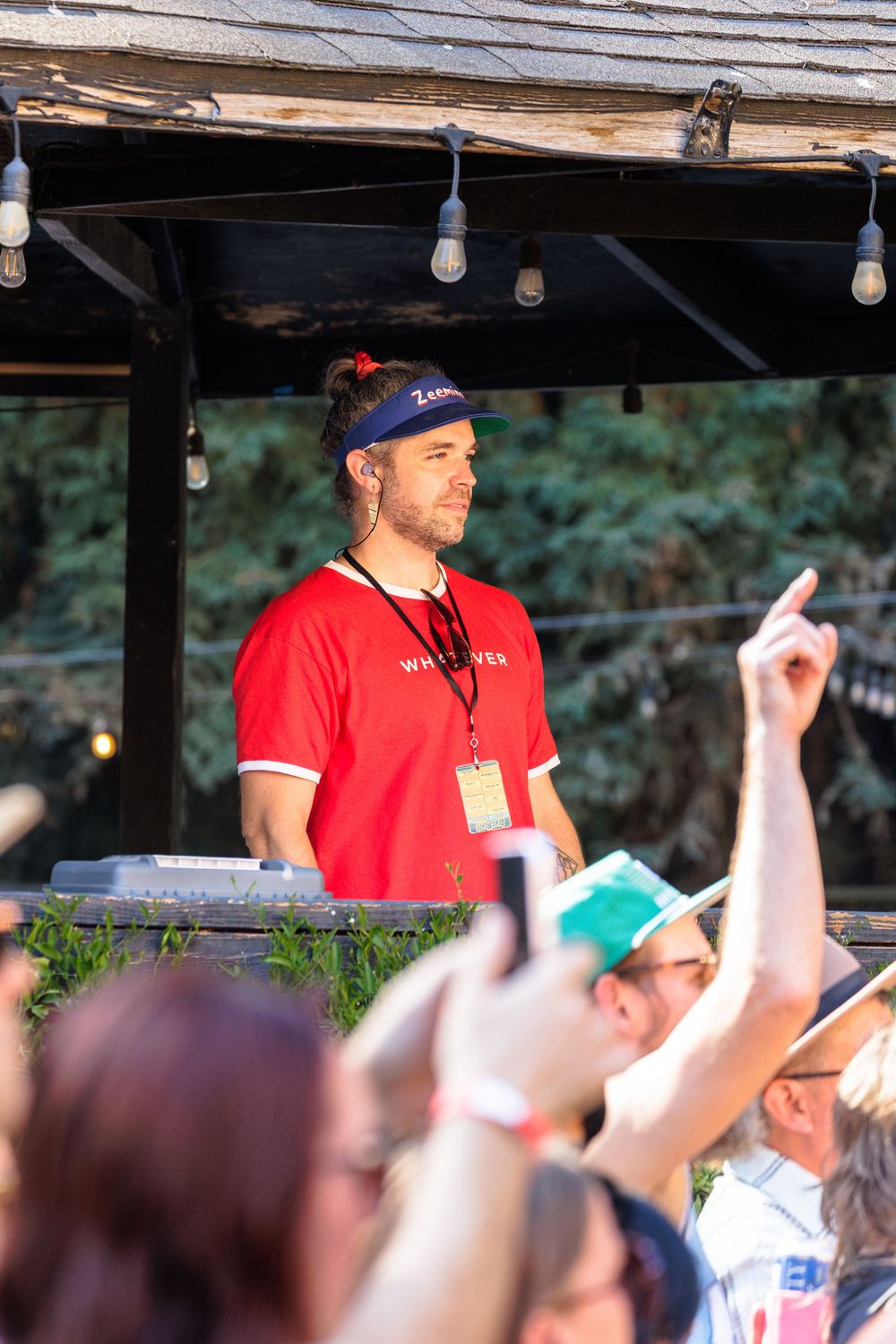 DJ in red shirt and blue cap performing at outdoor festival stage with crowd cheering in foreground.