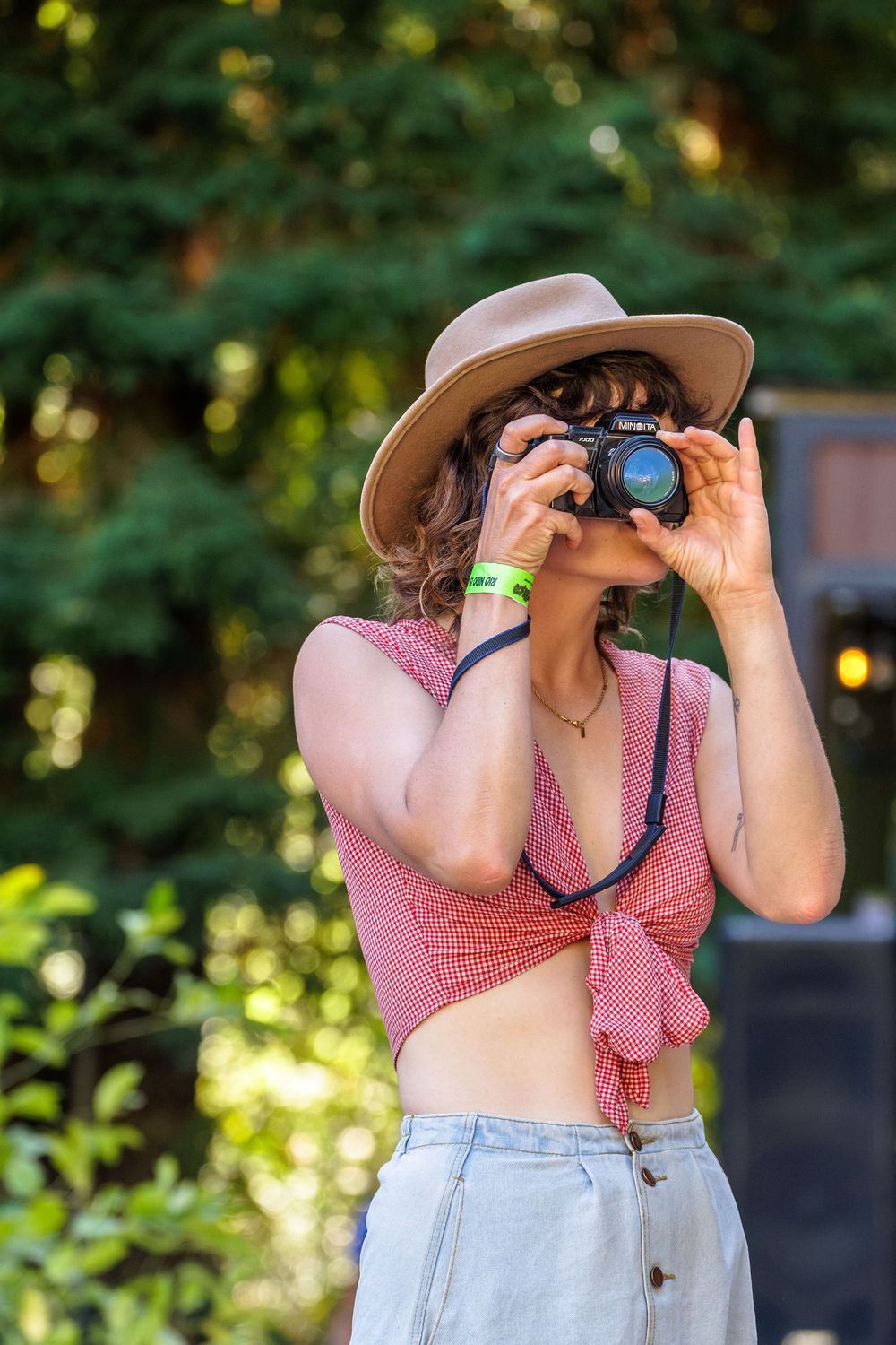 Young woman in tan hat and red crop top photographing with DSLR camera outdoors in green garden setting.