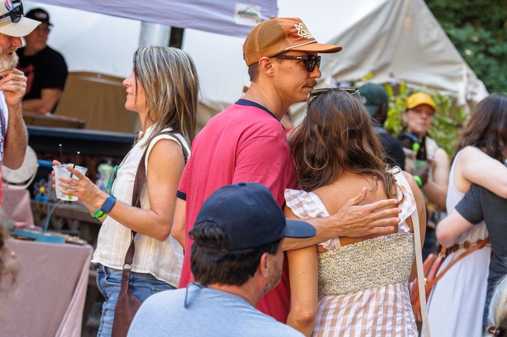 People socializing at an outdoor festival, with a man in a red shirt hugging a woman in a floral dress.