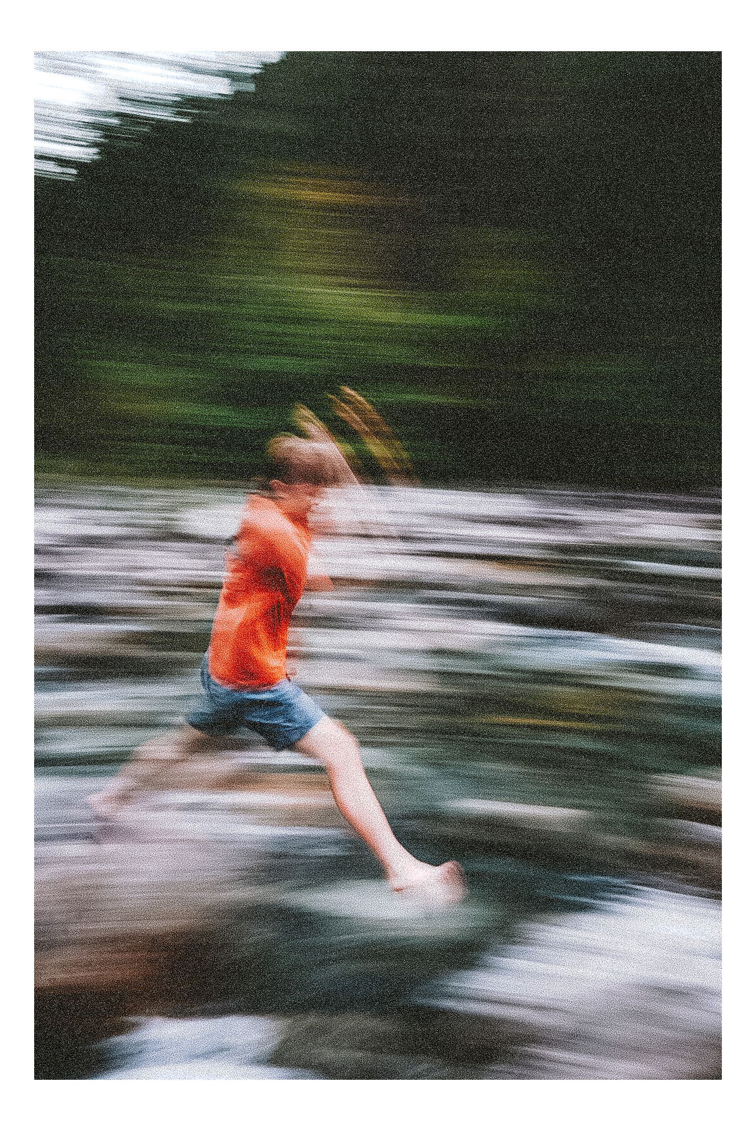 A blurred motion photo of a child in an orange shirt leaping over a rocky stream outdoors.