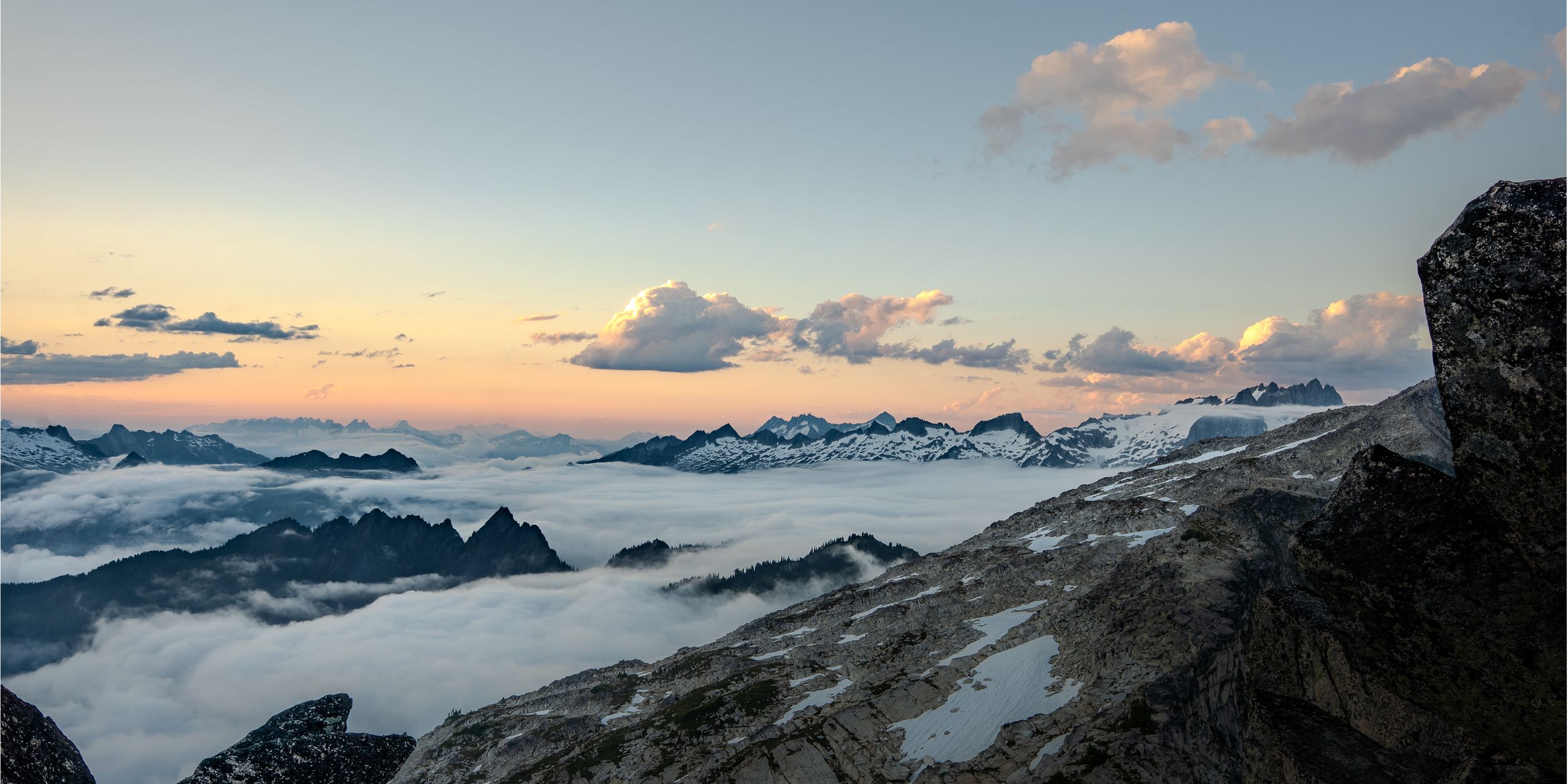 Panoramic mountain vista at sunset with peaks rising above a sea of clouds and snow-capped ridges glowing in warm light.