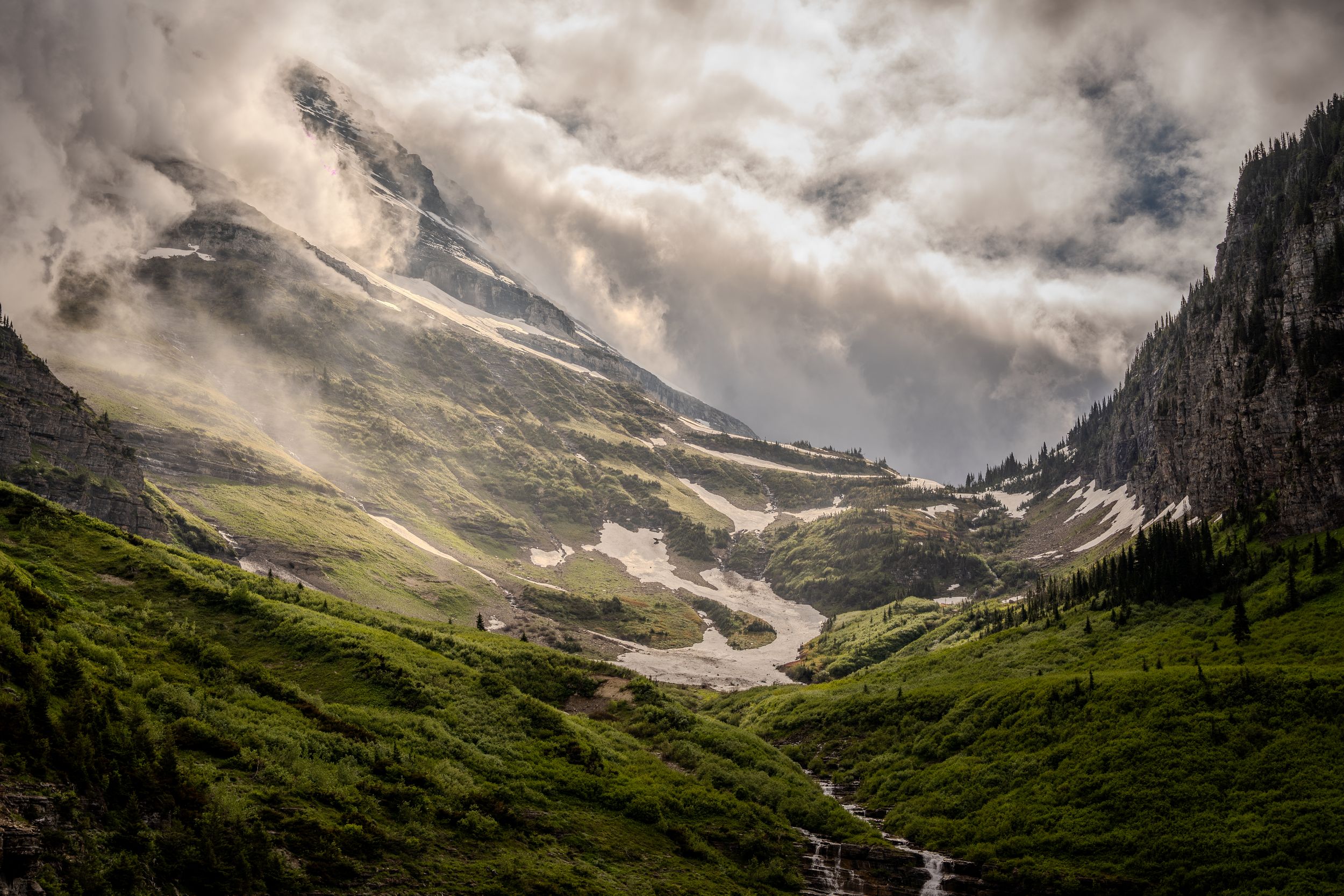 Dramatic alpine valley with lush green slopes, rocky peaks, and swirling storm clouds in a mountainous landscape.