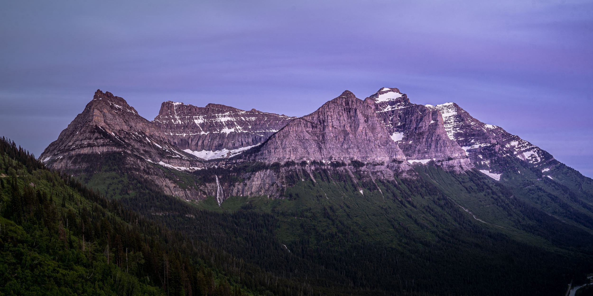Panoramic view of rugged mountain peaks with snow patches under a purple twilight sky in Glacier National Park.