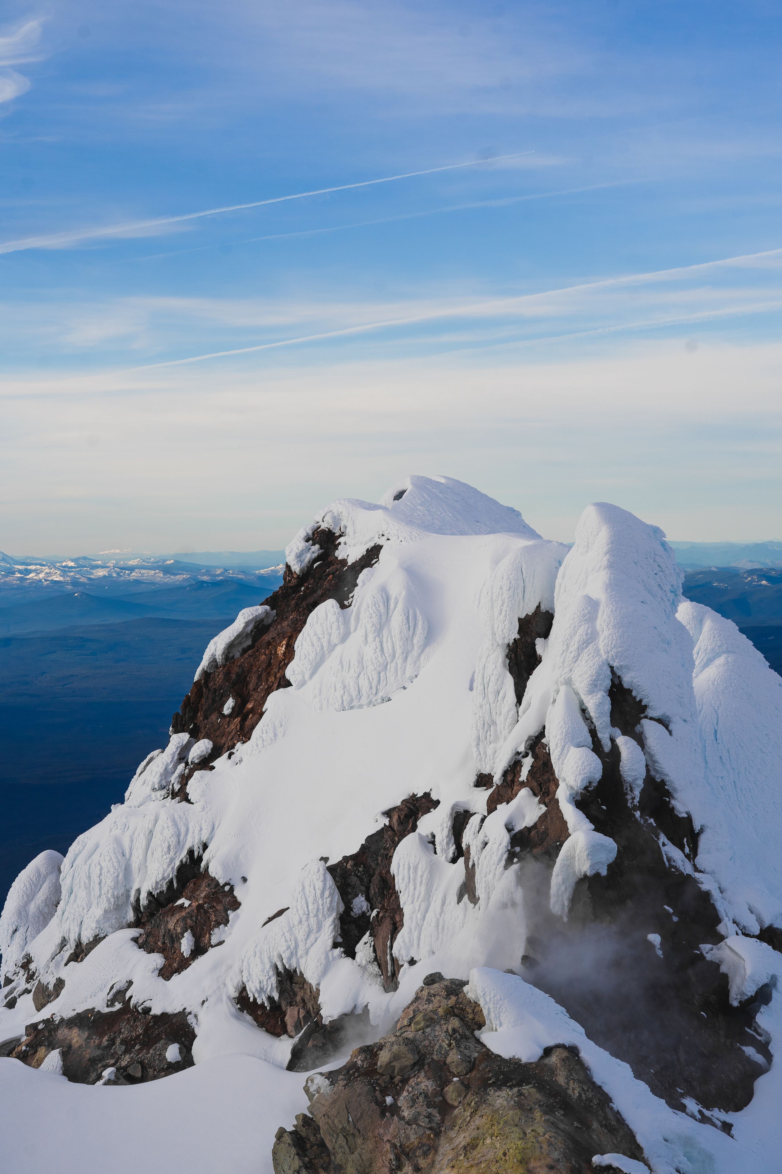 Snow-covered rocky mountain peak under a blue sky with wispy clouds and distant mountain ranges visible on the horizon.