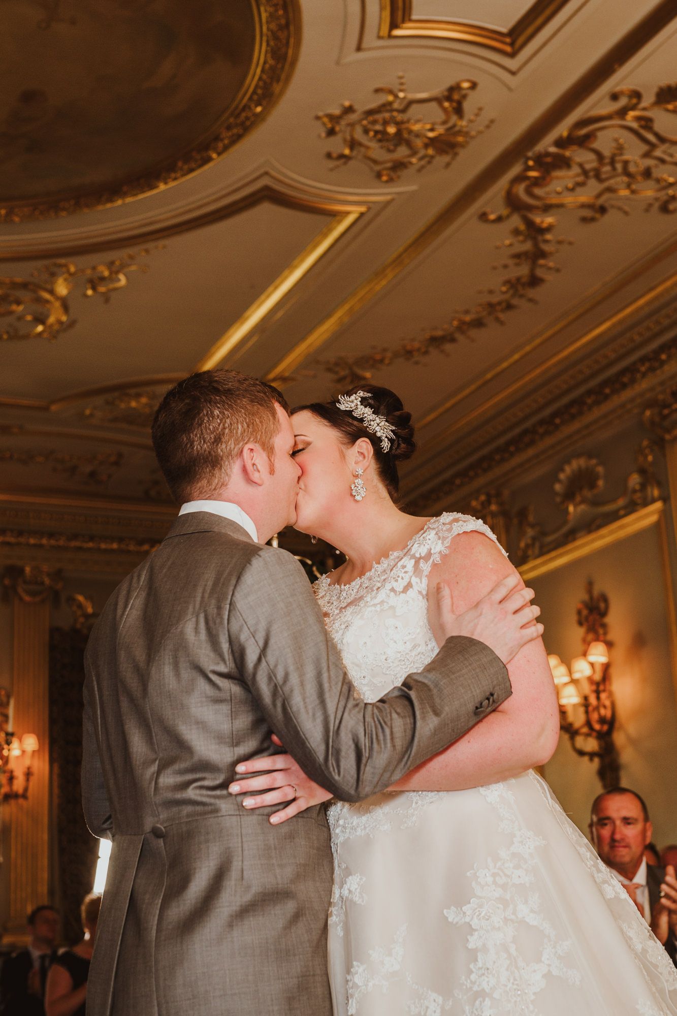 Bride and groom share their first kiss in an ornate golden ballroom during their elegant wedding ceremony.