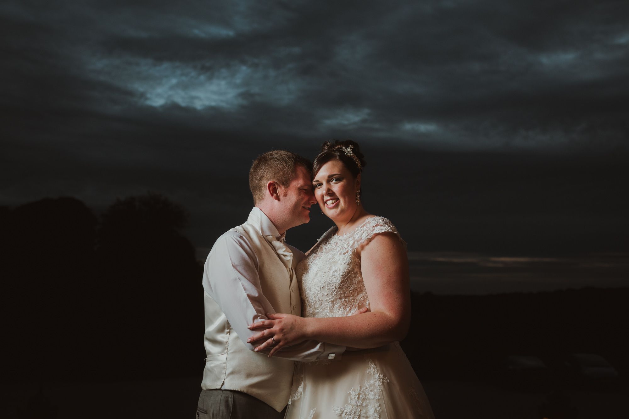 Couple embracing outdoors at dusk with dramatic stormy sky, bride in lace gown and groom in gray suit smiling together.