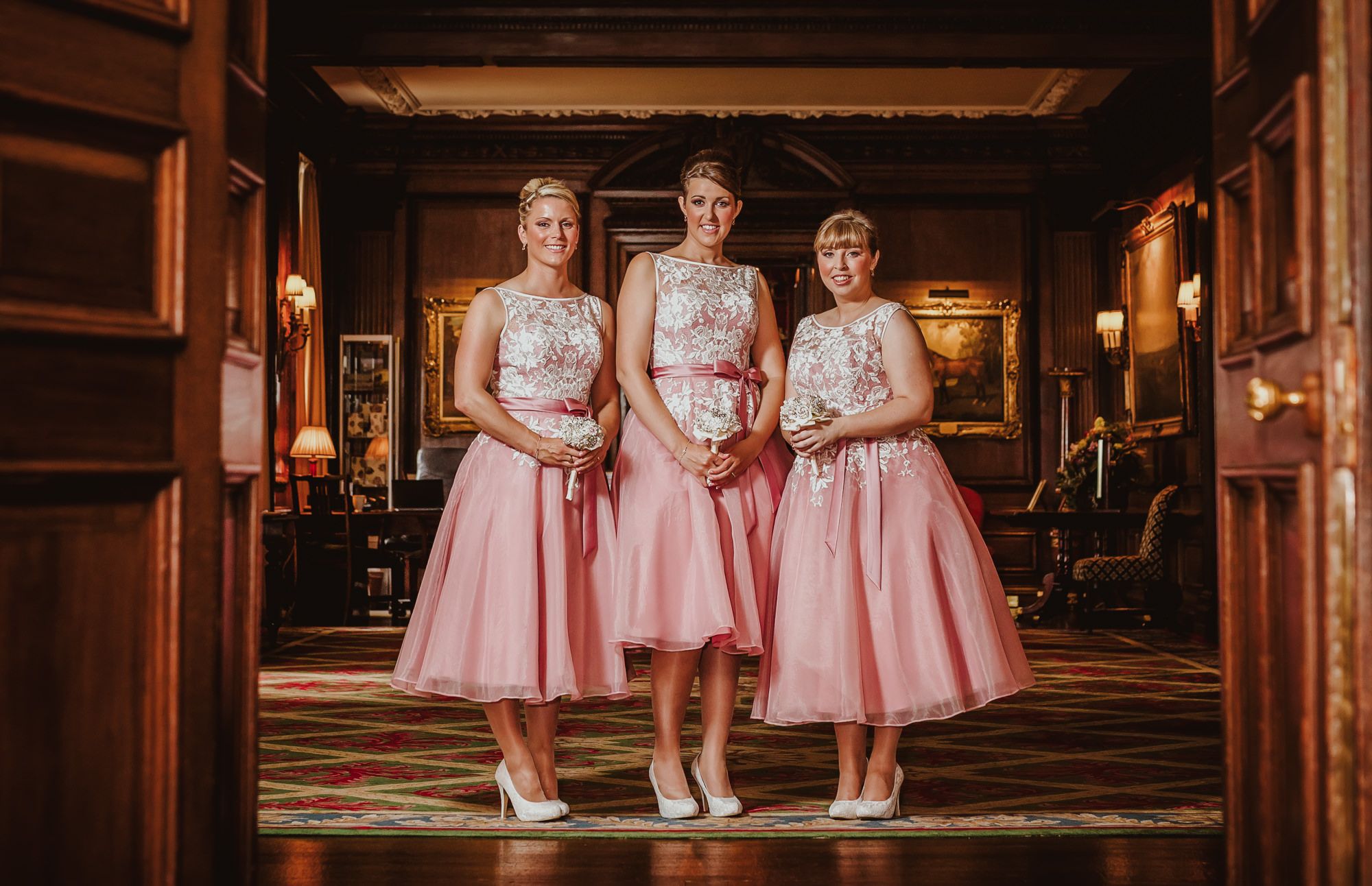 Three bridesmaids in matching pink tea-length dresses with lace bodices stand elegantly in a grand wooden hallway.