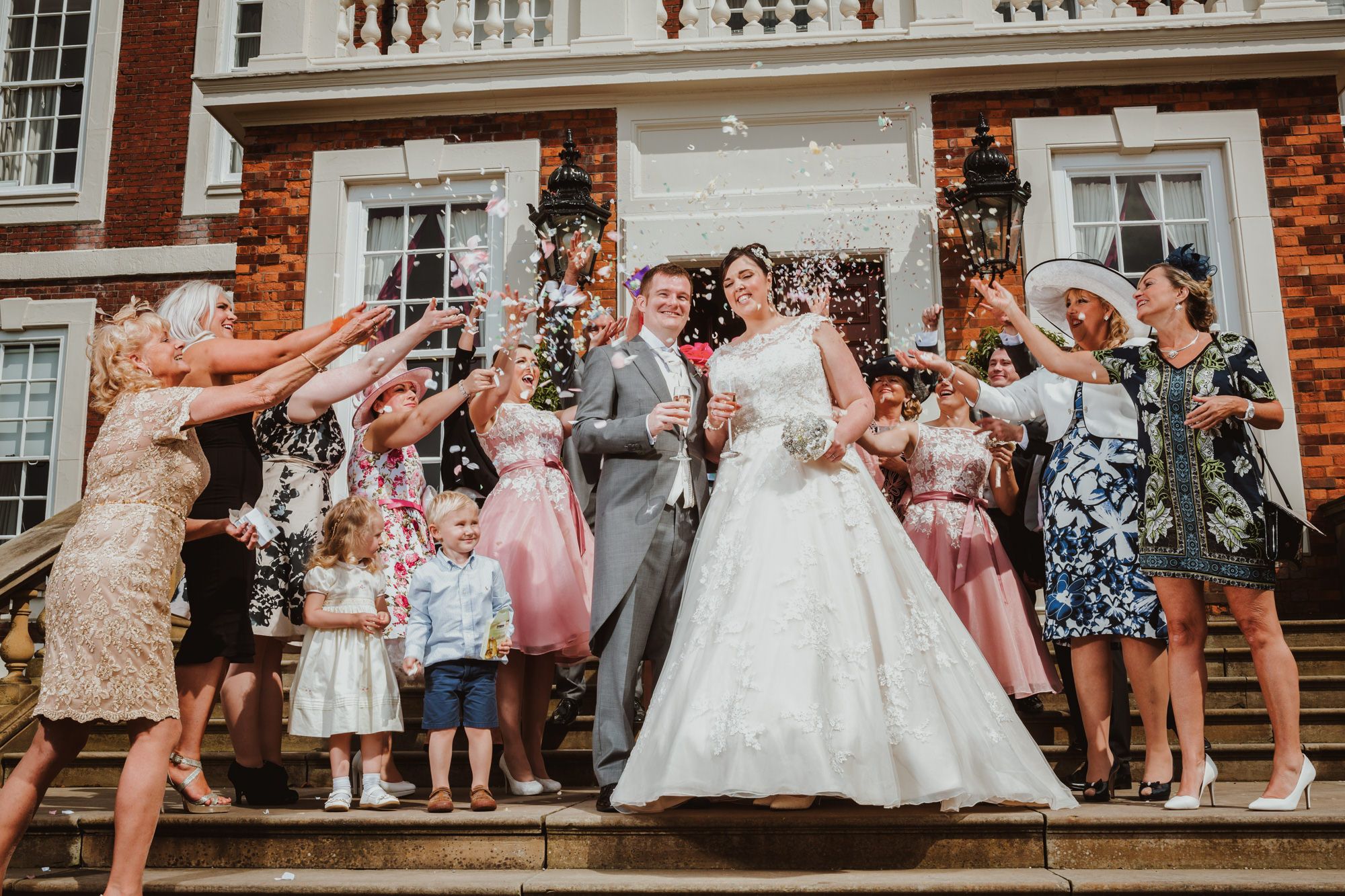 Bride and groom celebrate on steps with guests throwing confetti at elegant red brick wedding venue.