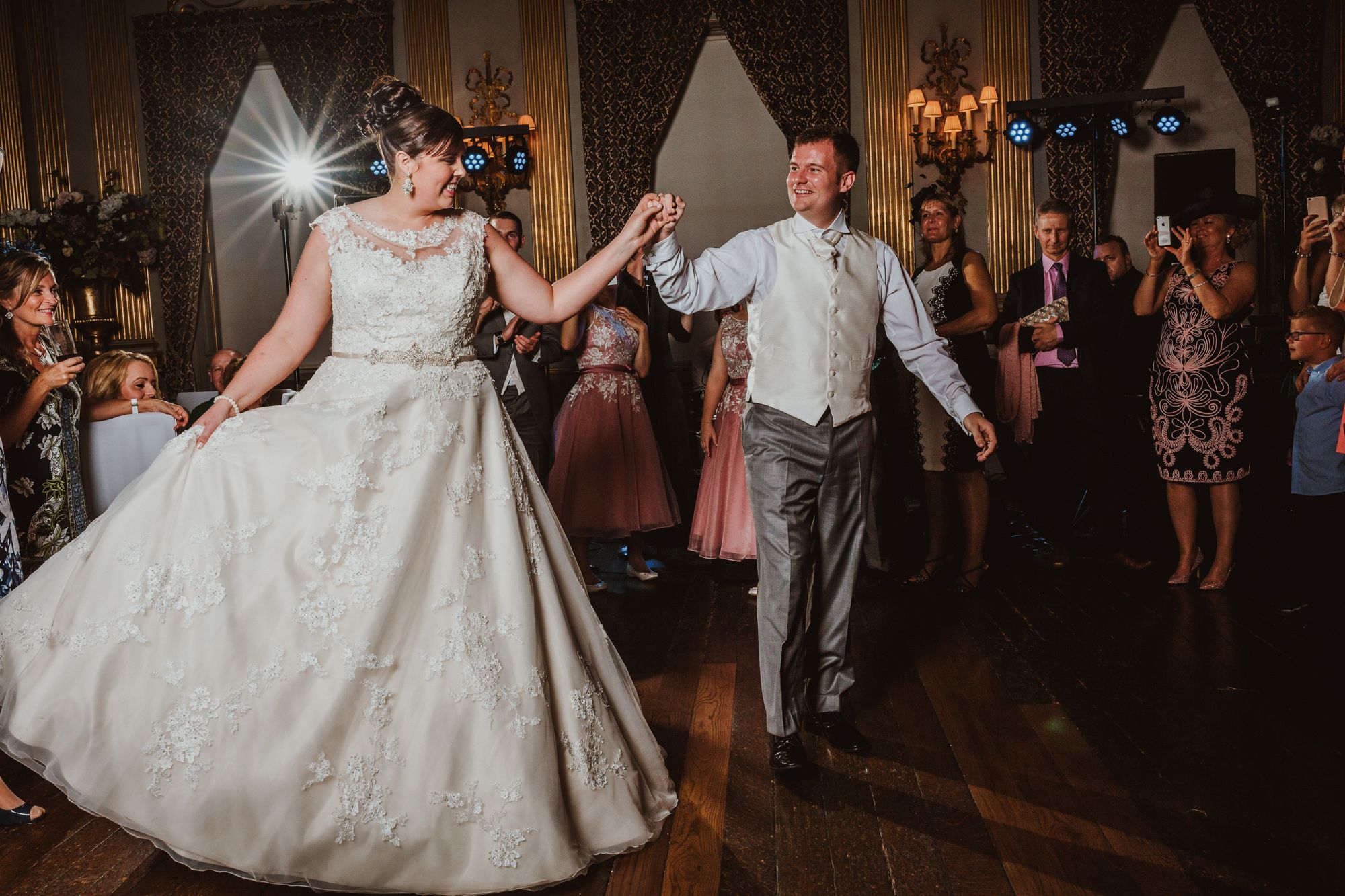 Bride and groom share first dance at elegant wedding reception, bride in lace ballgown, guests celebrating in background.