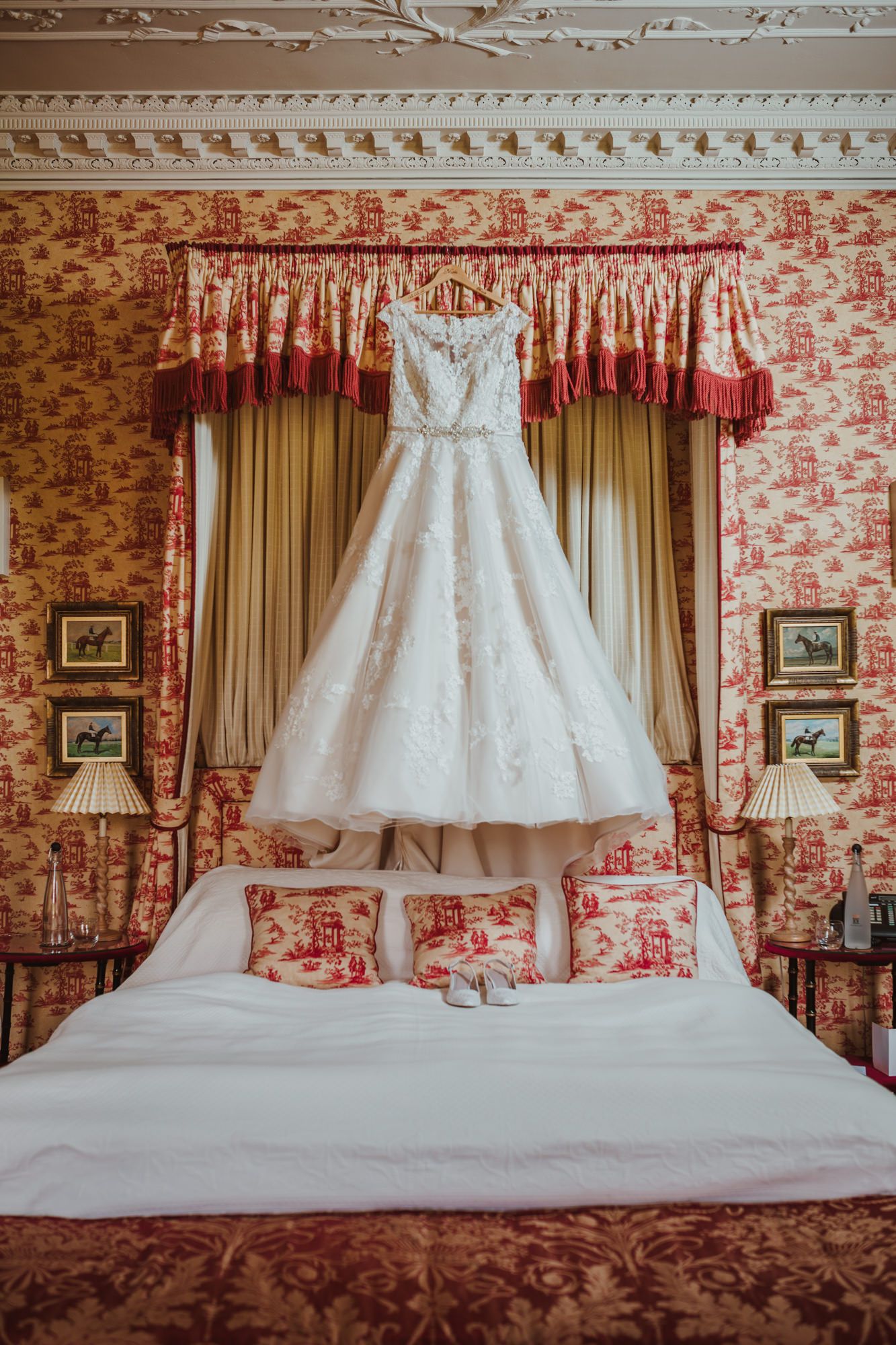 White lace ball gown wedding dress hanging over a bed in an elegant red toile de jouy decorated bedroom.