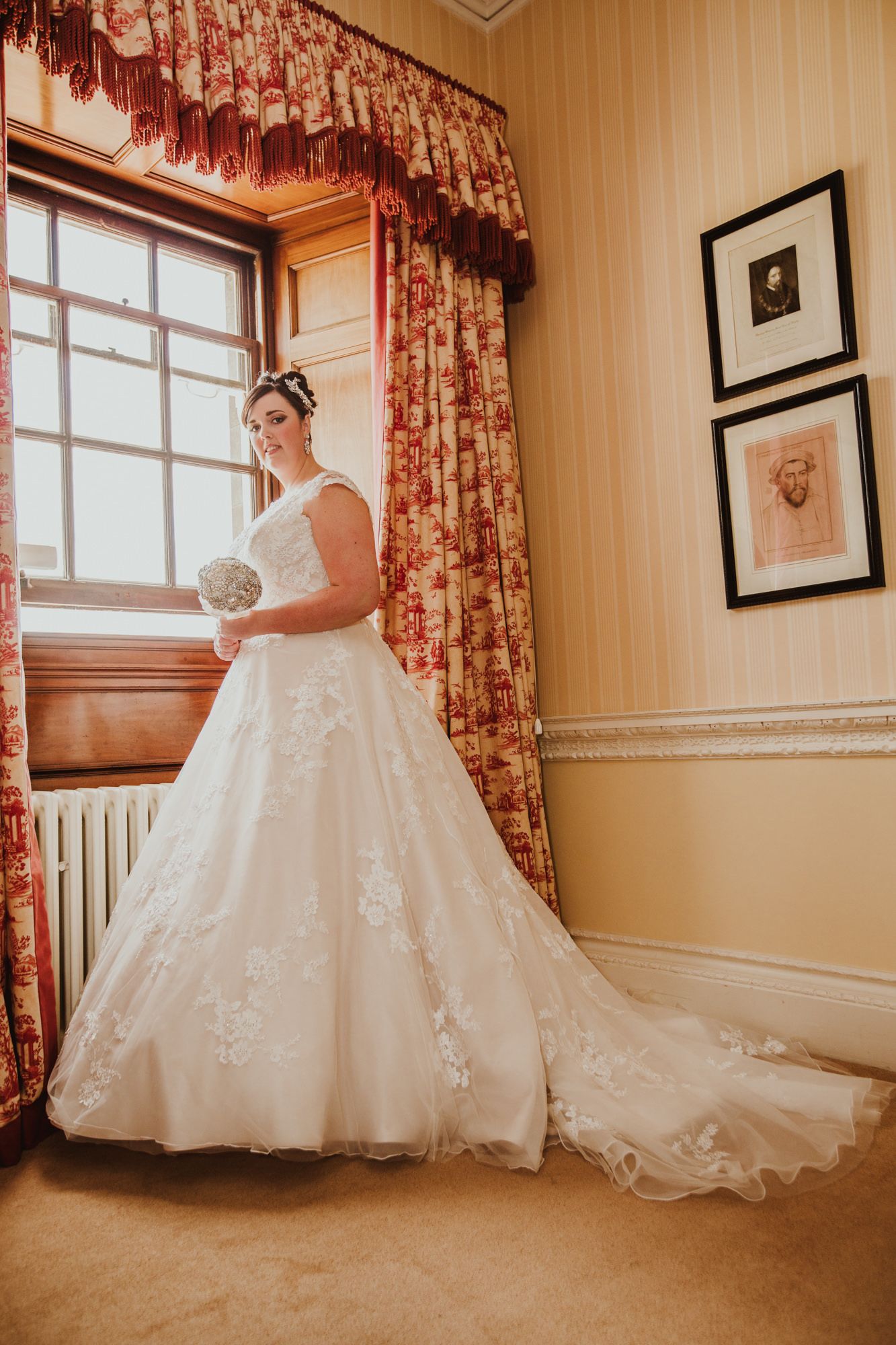 Bride in white lace ball gown wedding dress standing by window holding bouquet in elegant room with floral curtains.