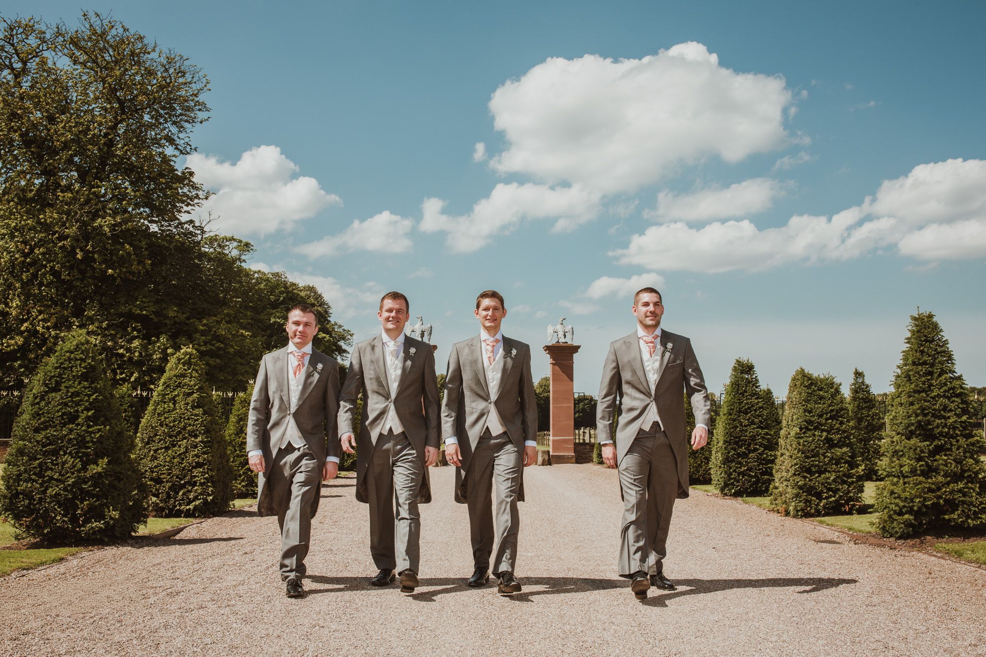 Four groomsmen in grey suits walking confidently down a gravel path surrounded by manicured hedges on a sunny wedding day.