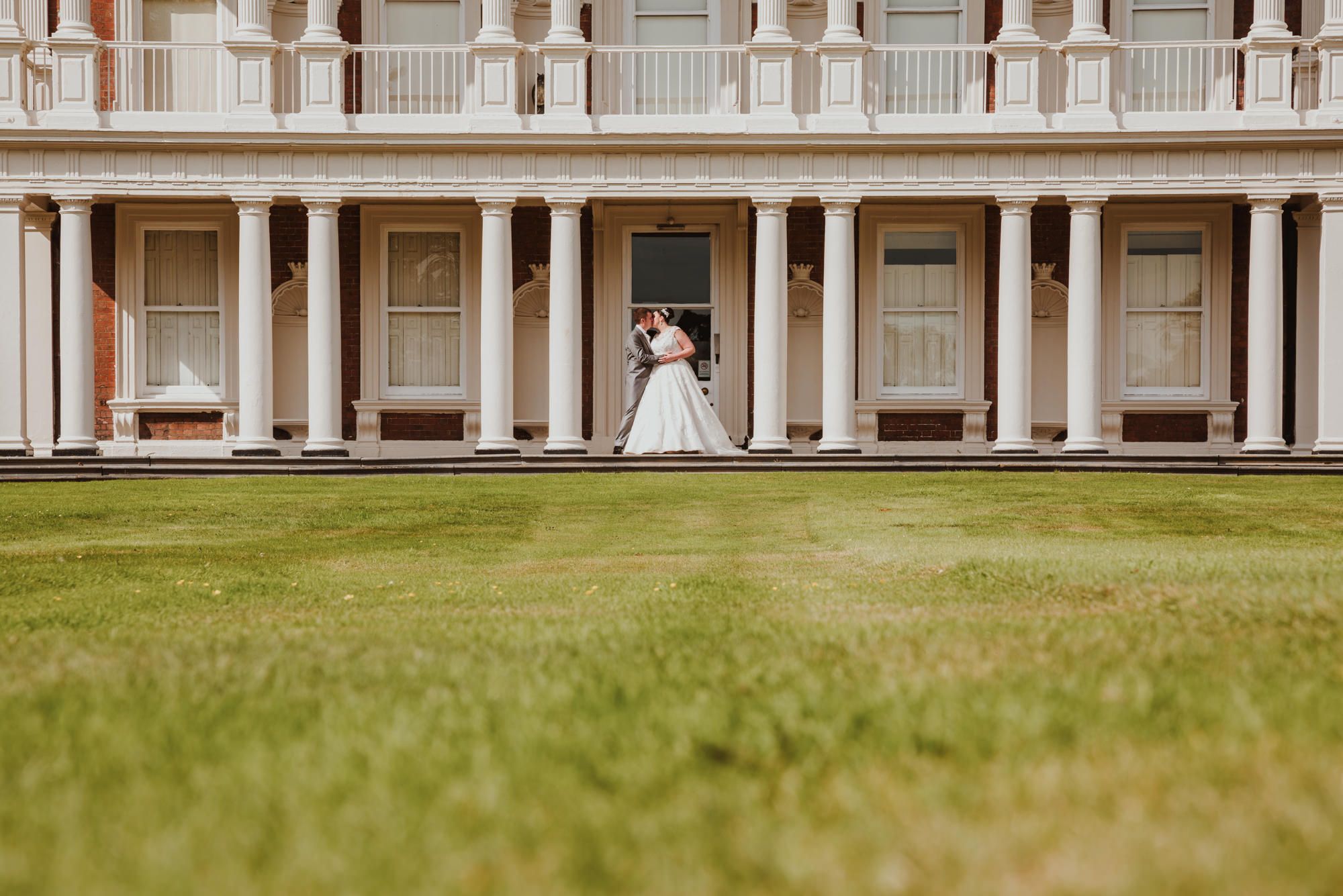 Bride and groom share a romantic kiss on lush green lawn before elegant white colonial mansion with tall columns.