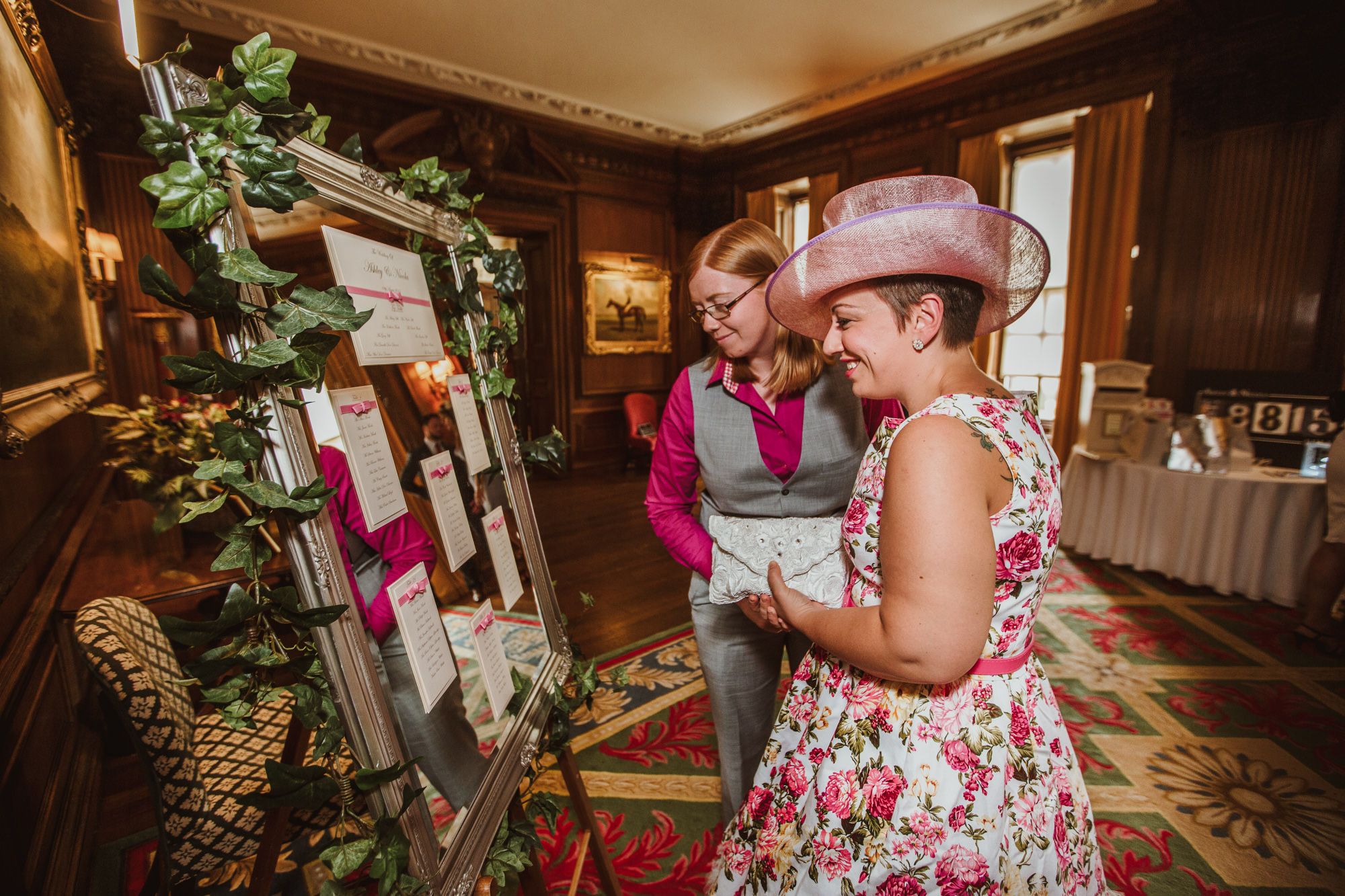 Two women in elegant attire review a floral wedding seating chart display in a grand venue.