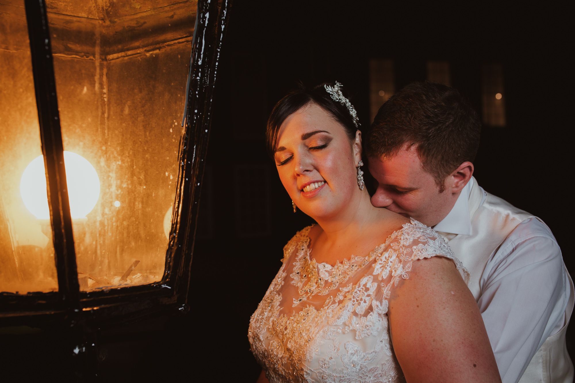 Bride and groom share an intimate moment beside a glowing lantern, bride in lace wedding gown with hair accessory.