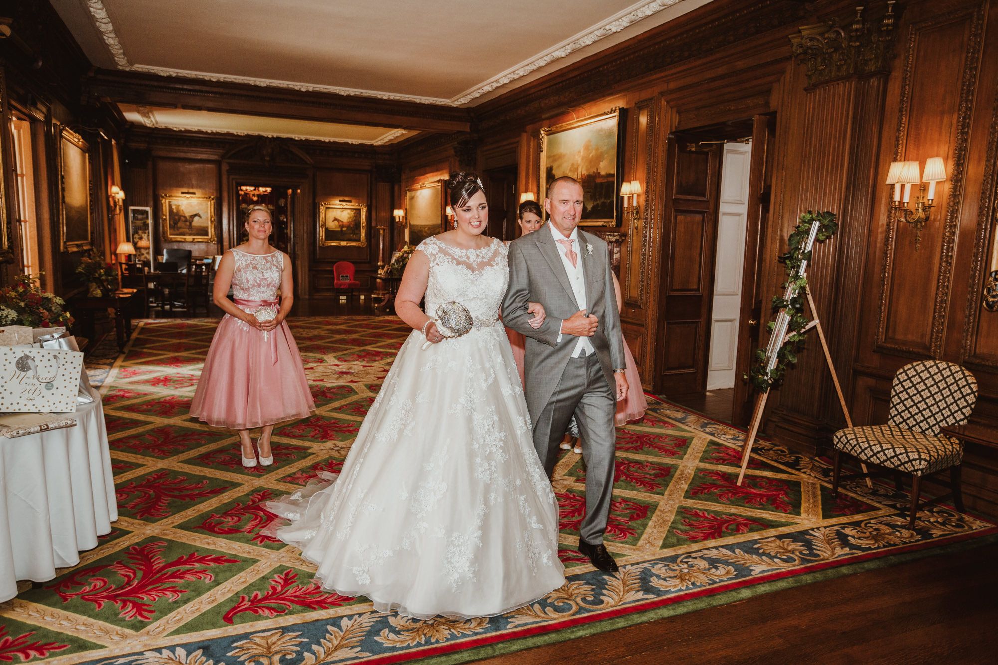 Bride and groom walk through ornate wood-paneled wedding venue as guests watch in background.
