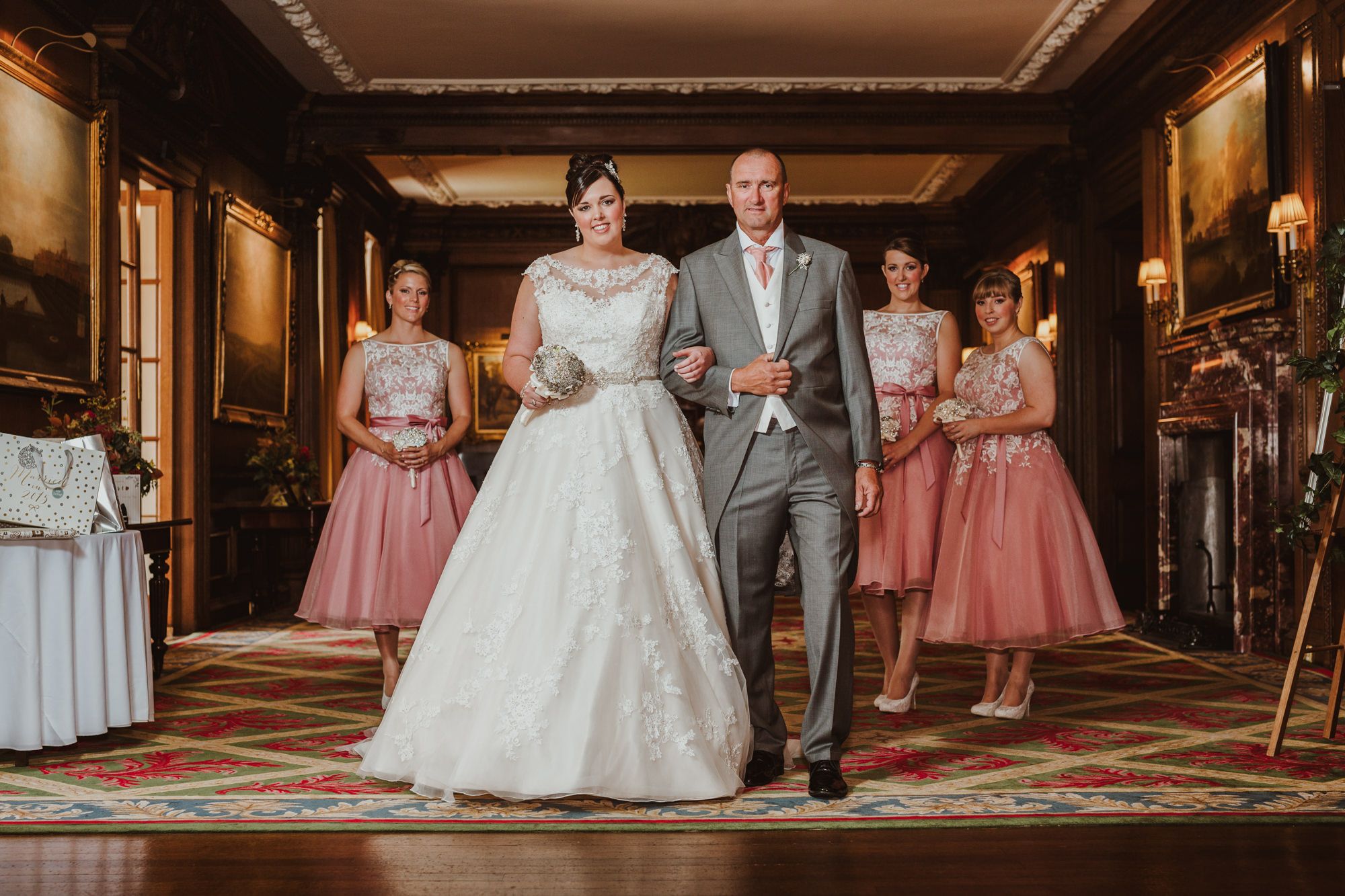 Bride in white gown walks with groom in gray suit, bridesmaids in pink dresses behind them in elegant wood-paneled hall.