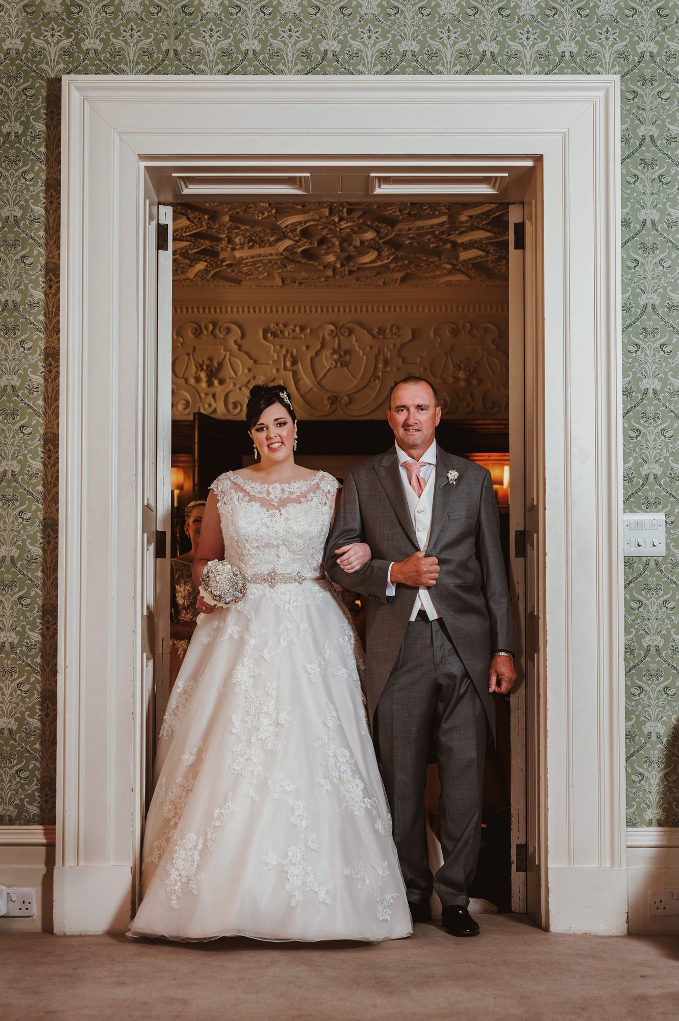 Bride in lace gown and groom in gray suit walk through ornate doorway at elegant wedding venue.