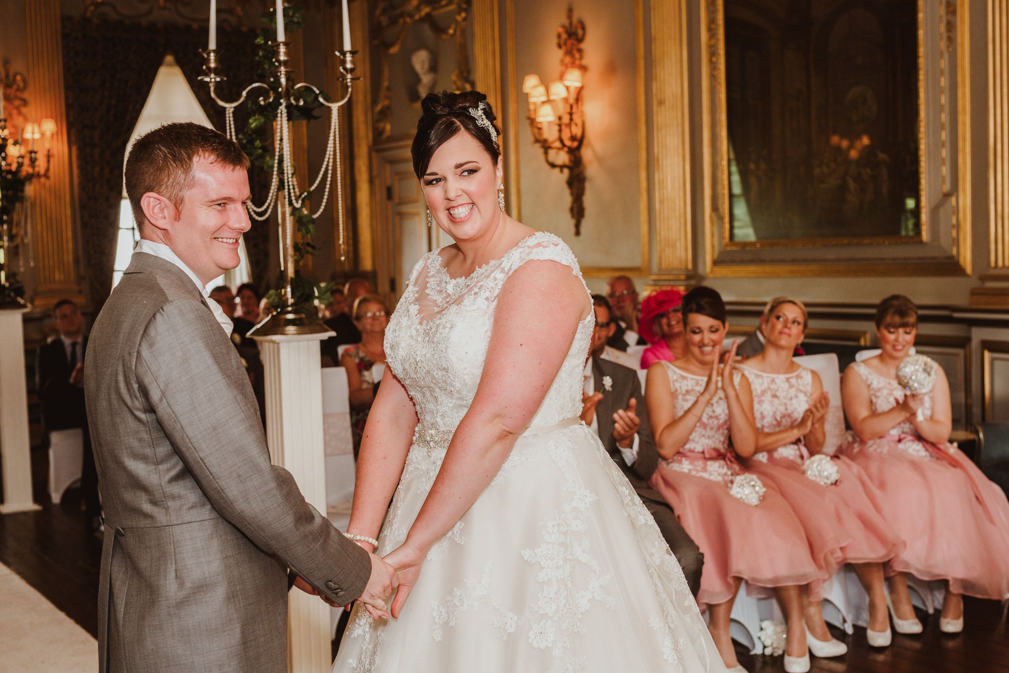 Bride and groom holding hands during wedding ceremony, bridesmaids in pink dresses seated in elegant venue.