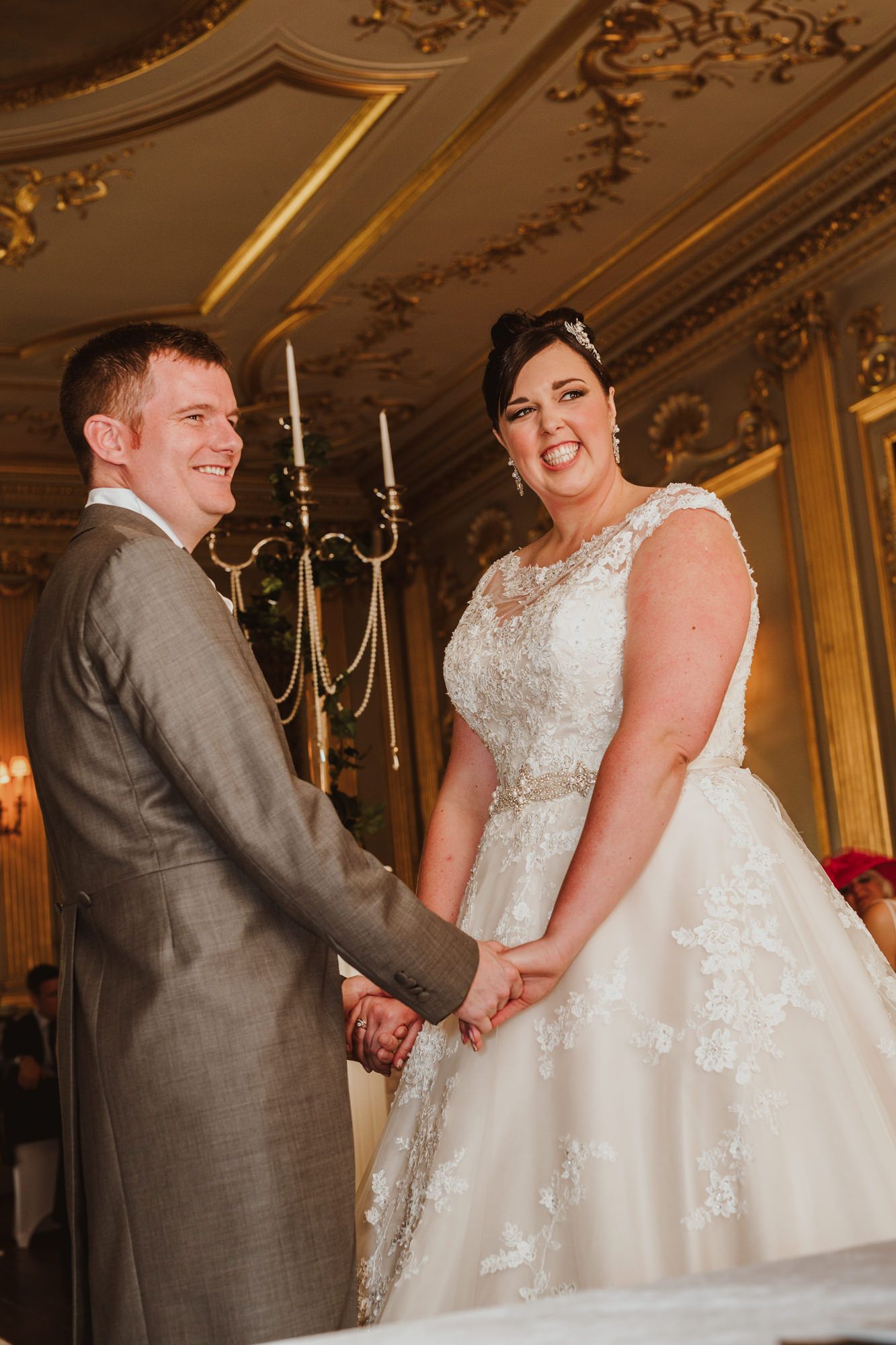 Smiling bride and groom hold hands at ornate gold wedding ceremony altar.