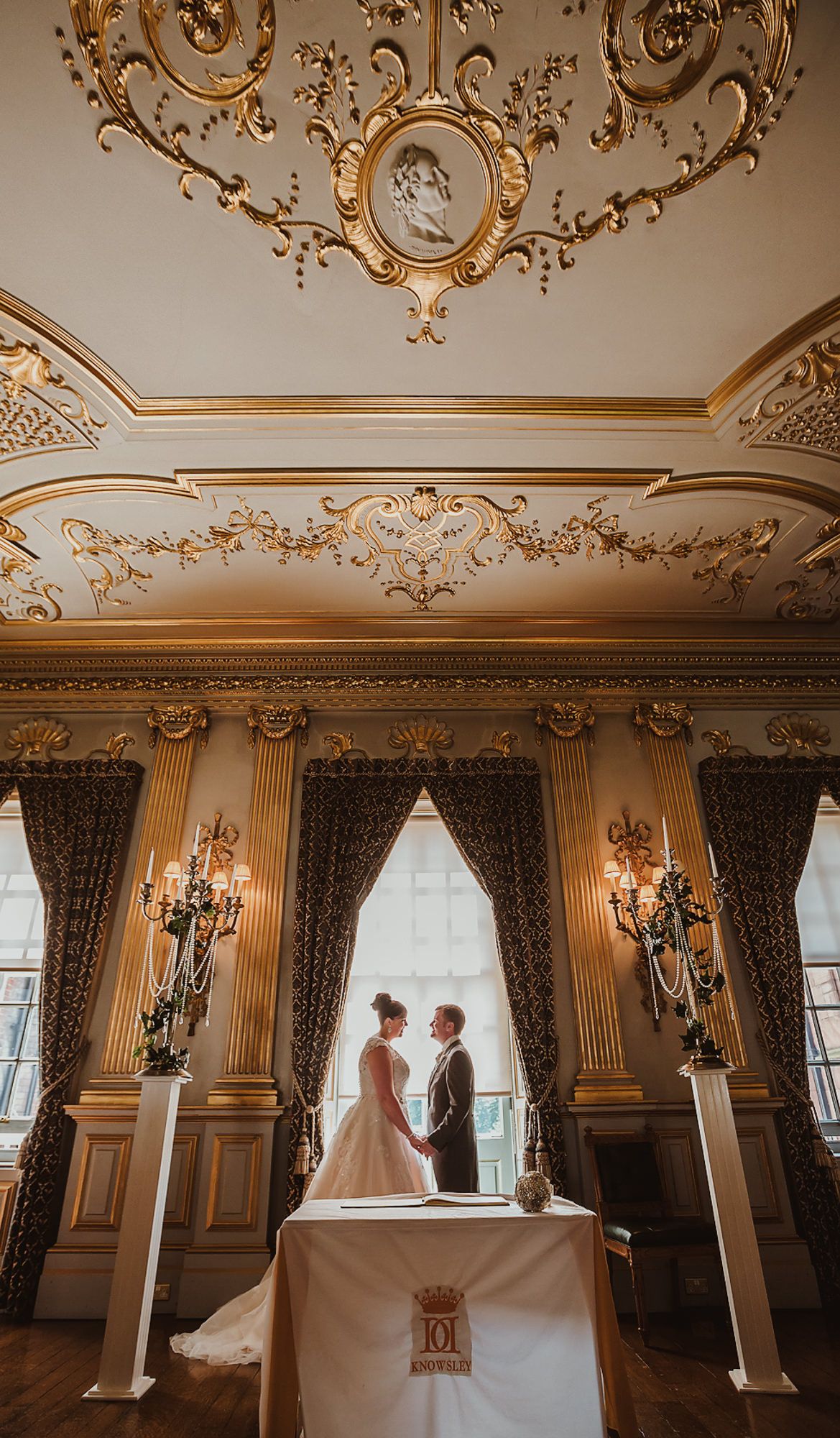 Bride and groom share a romantic moment in an ornate gold ballroom with baroque ceiling details and candlelit sconces.
