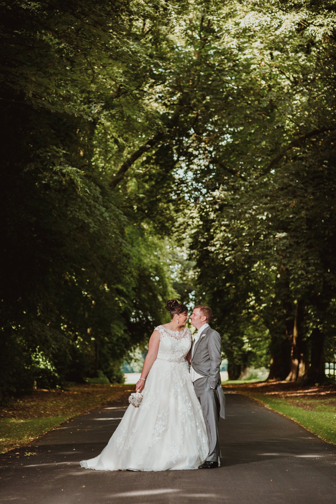 Bride and groom share a romantic kiss on a tree-lined path in lush green surroundings on their wedding day.