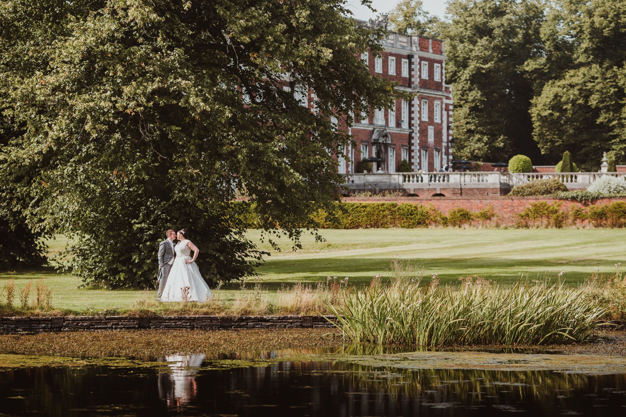 Bride and groom embrace by a tranquil pond with a grand red-brick manor house surrounded by lush gardens.