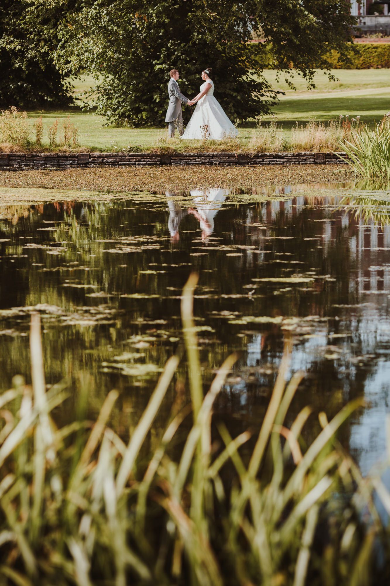 Bride and groom holding hands by a reflective pond with lily pads, surrounded by lush greenery on their wedding day.