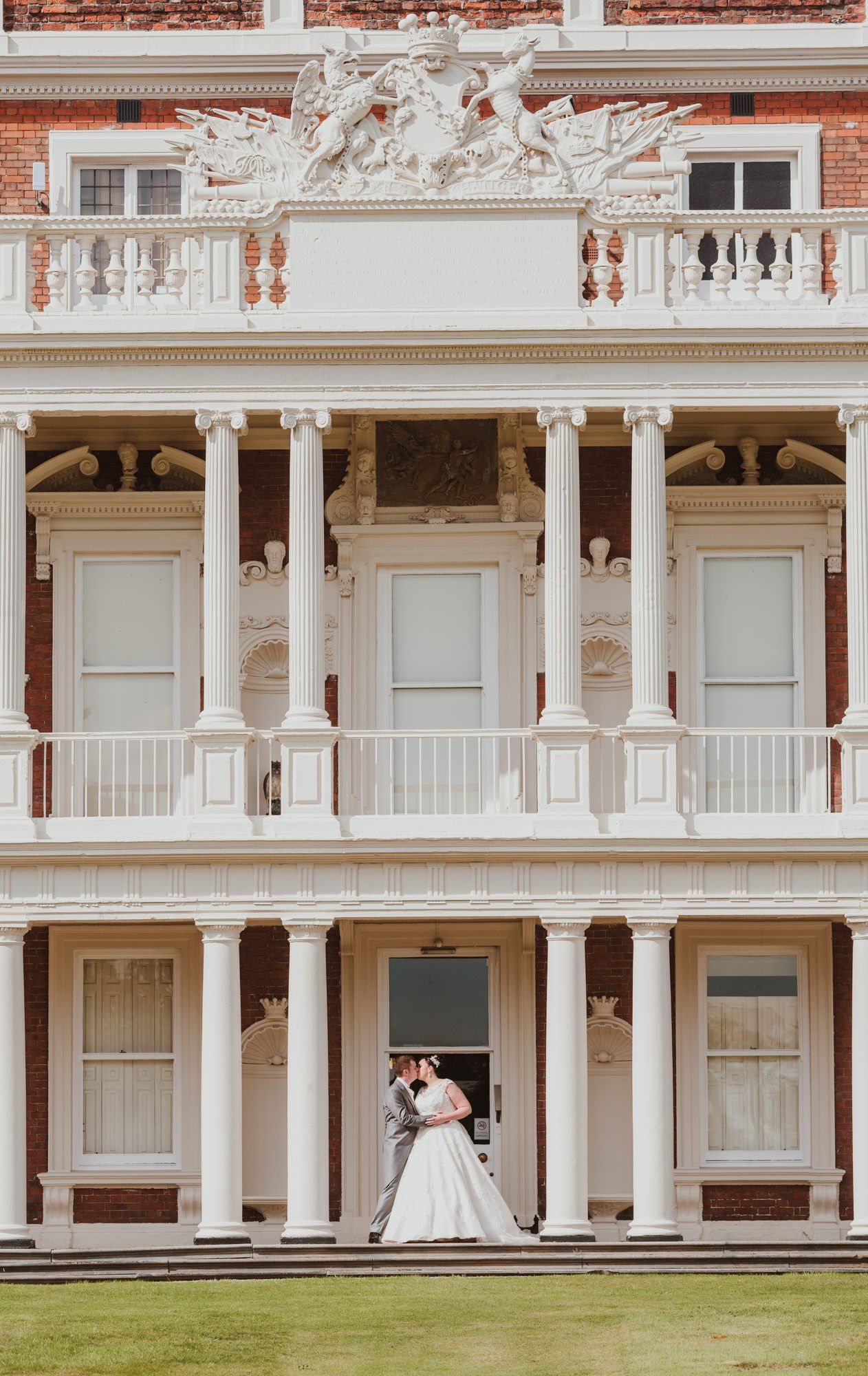 Bride and groom share a kiss on the grand portico of an elegant red brick mansion with white columns.