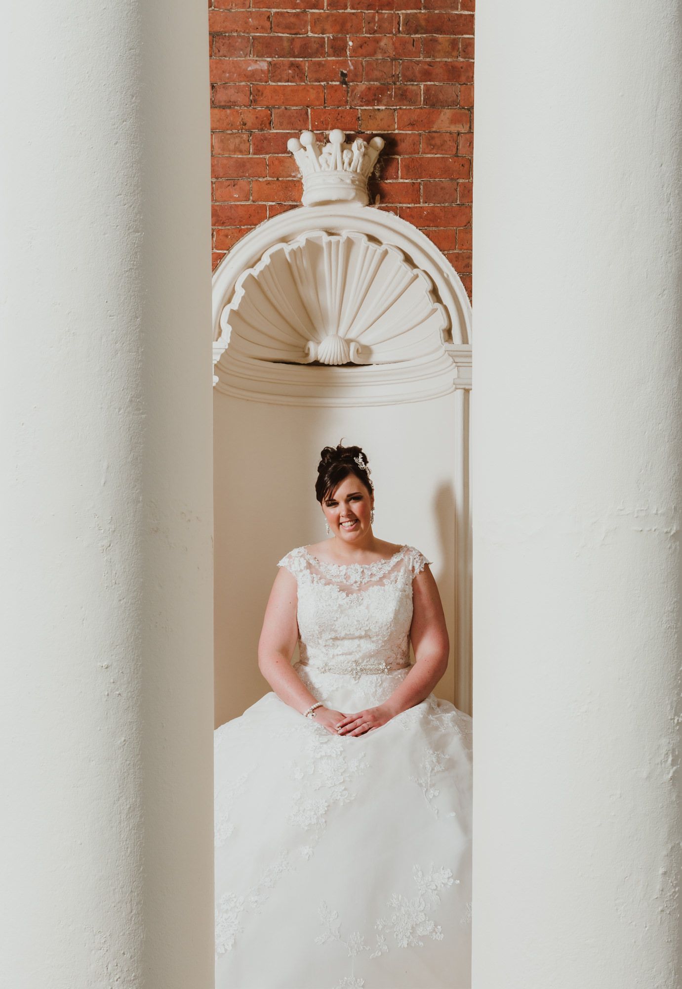 Bride in lace wedding gown posing between white columns with ornate architectural details and brick wall backdrop.