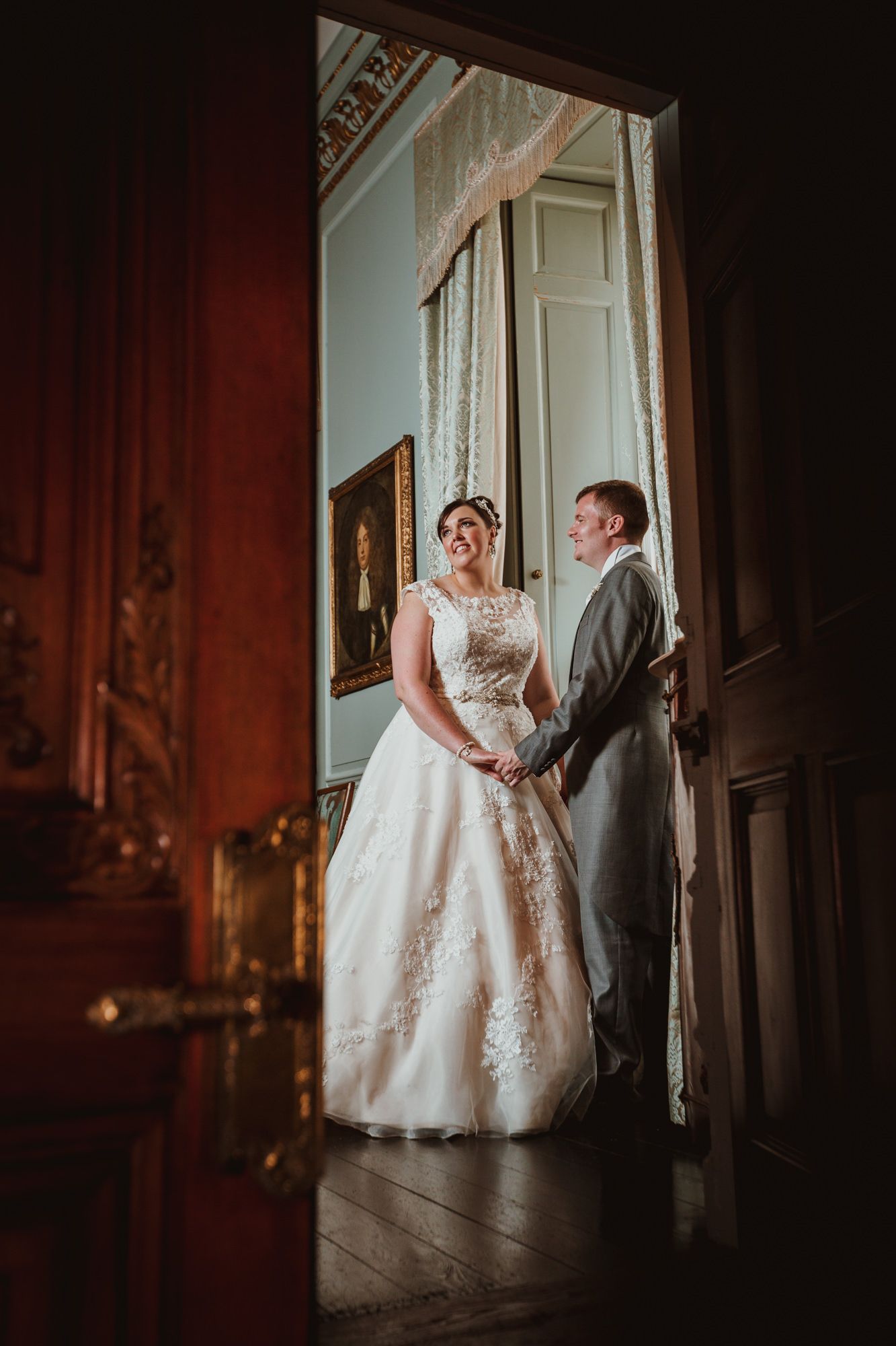 Bride and groom holding hands in elegant doorway of historic venue, bride wearing lace ballgown wedding dress.
