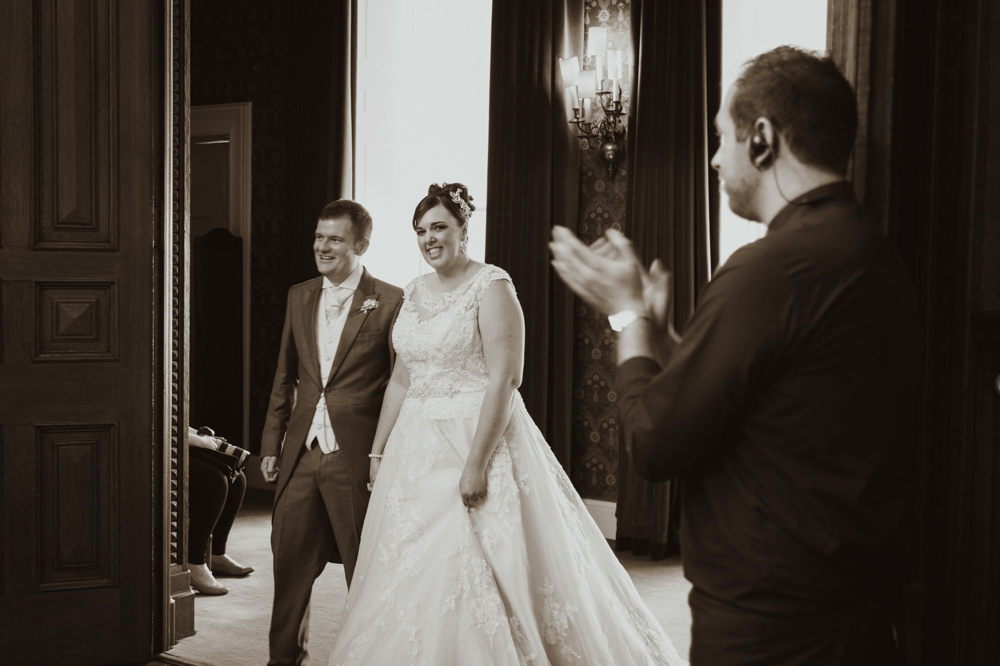 Bride in white gown walks with escort as guest applauds in elegant sepia-toned wedding venue.