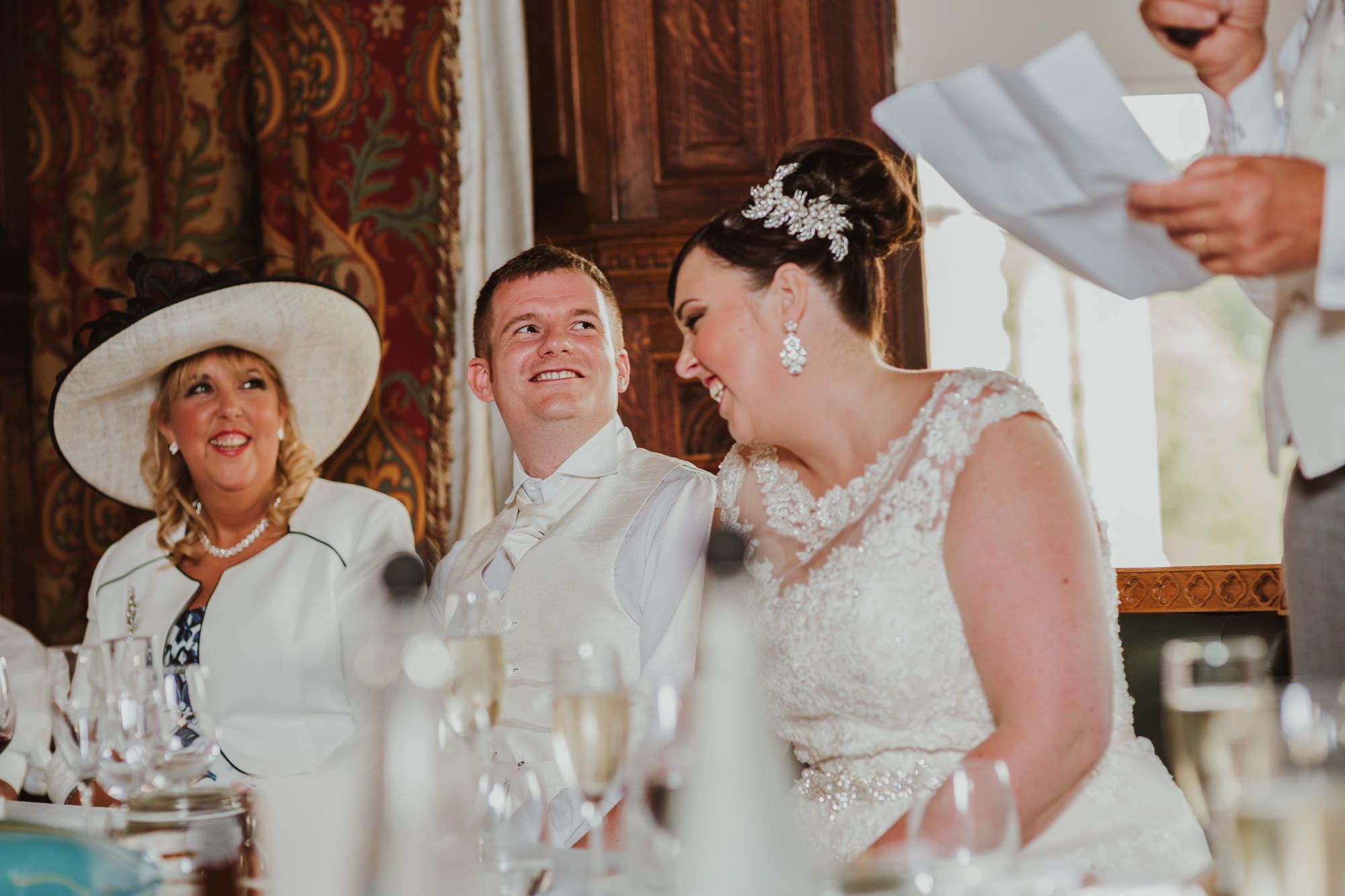Bride and groom laughing at wedding reception table with elegant decor and guest in white hat.