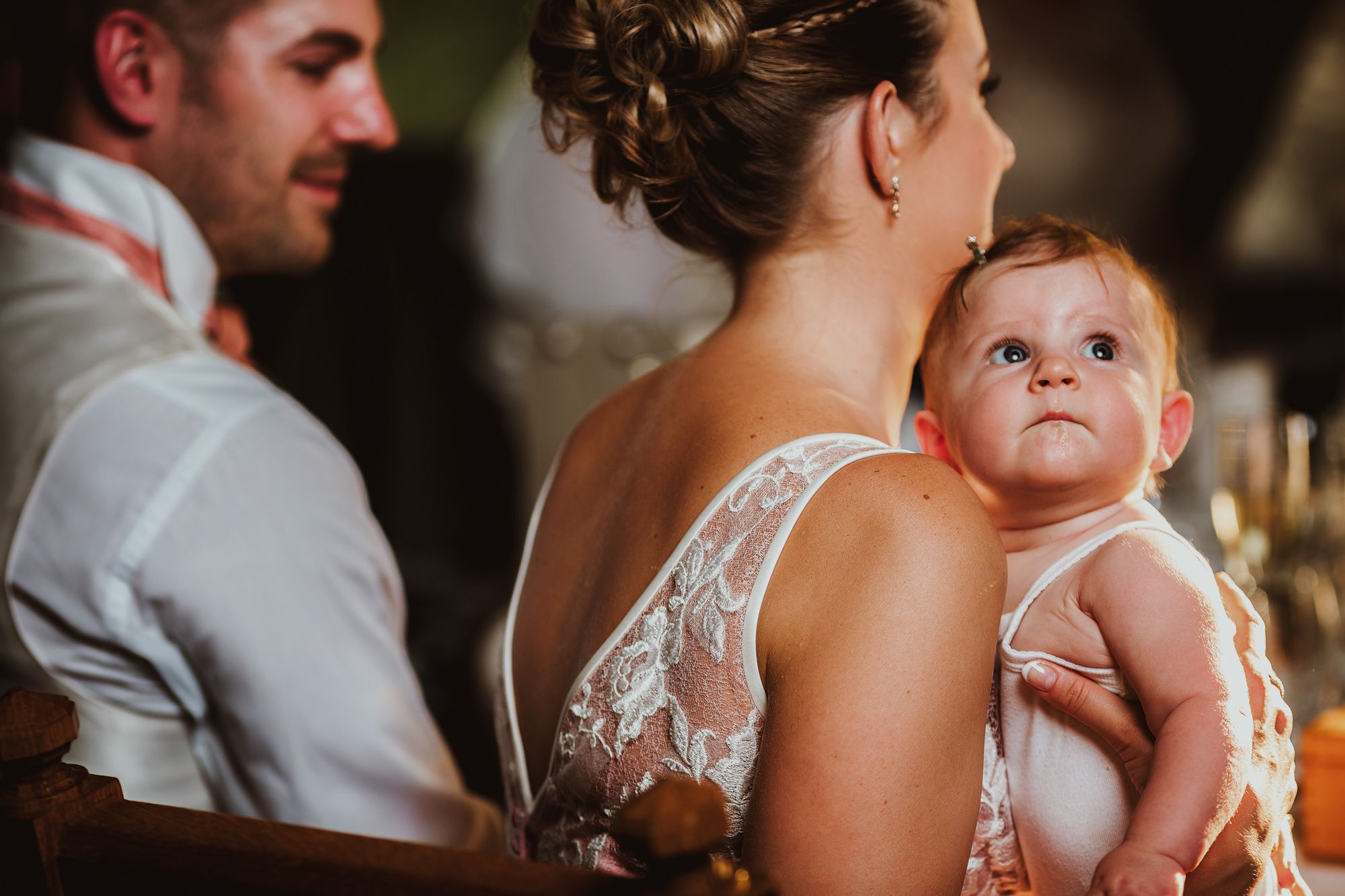 A bride in lace gown holds an adorable wide-eyed baby at a wedding reception, with groom seated beside her.
