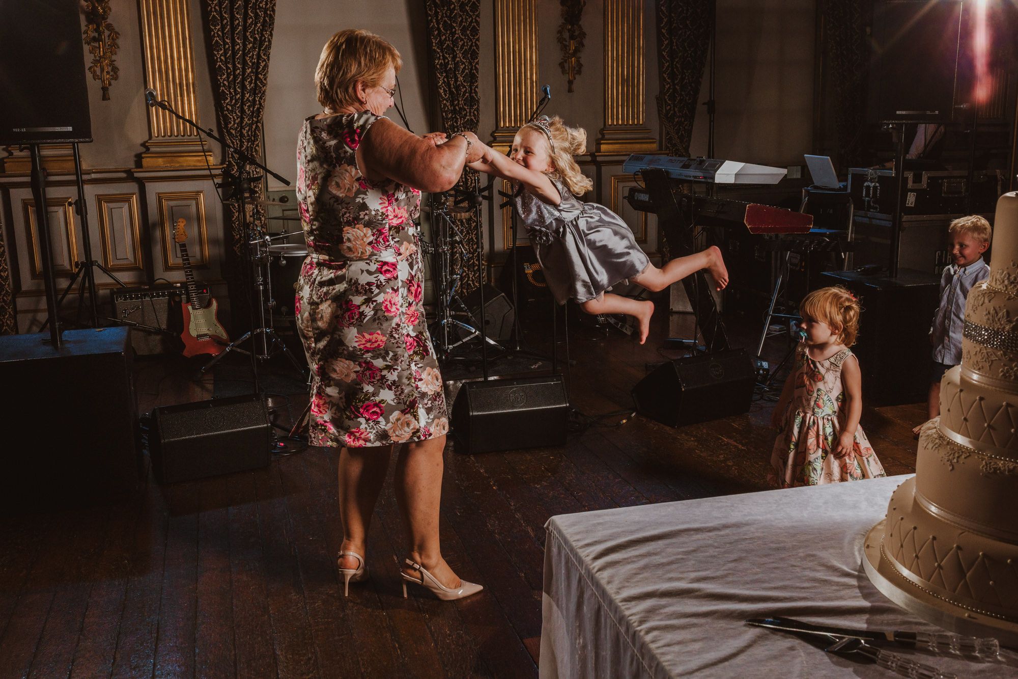Woman in floral dress playfully swinging a child at a wedding reception near a tiered cake.