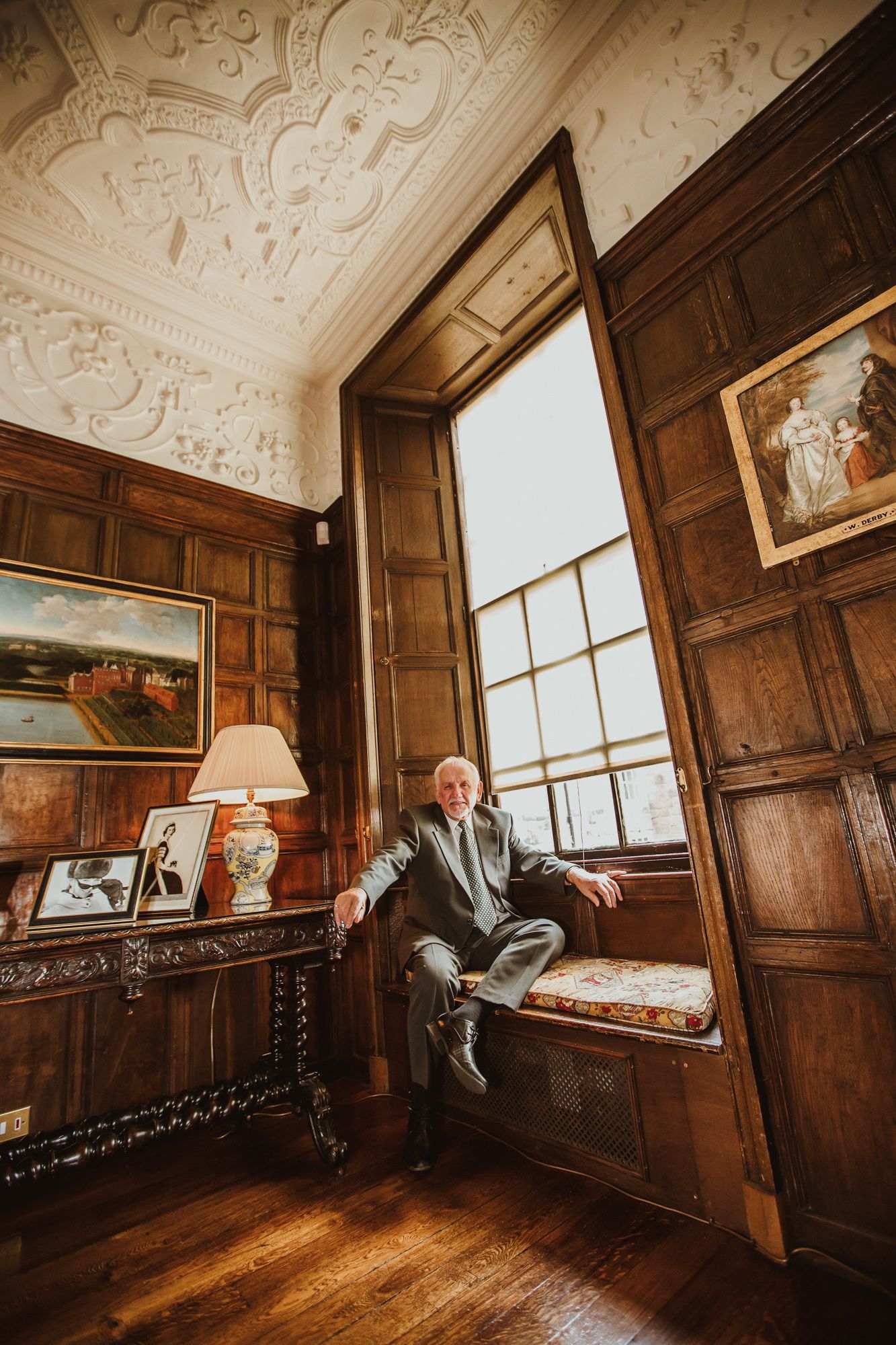 Elderly man sitting by a large window in an ornate wood-paneled room with decorative plaster ceiling.