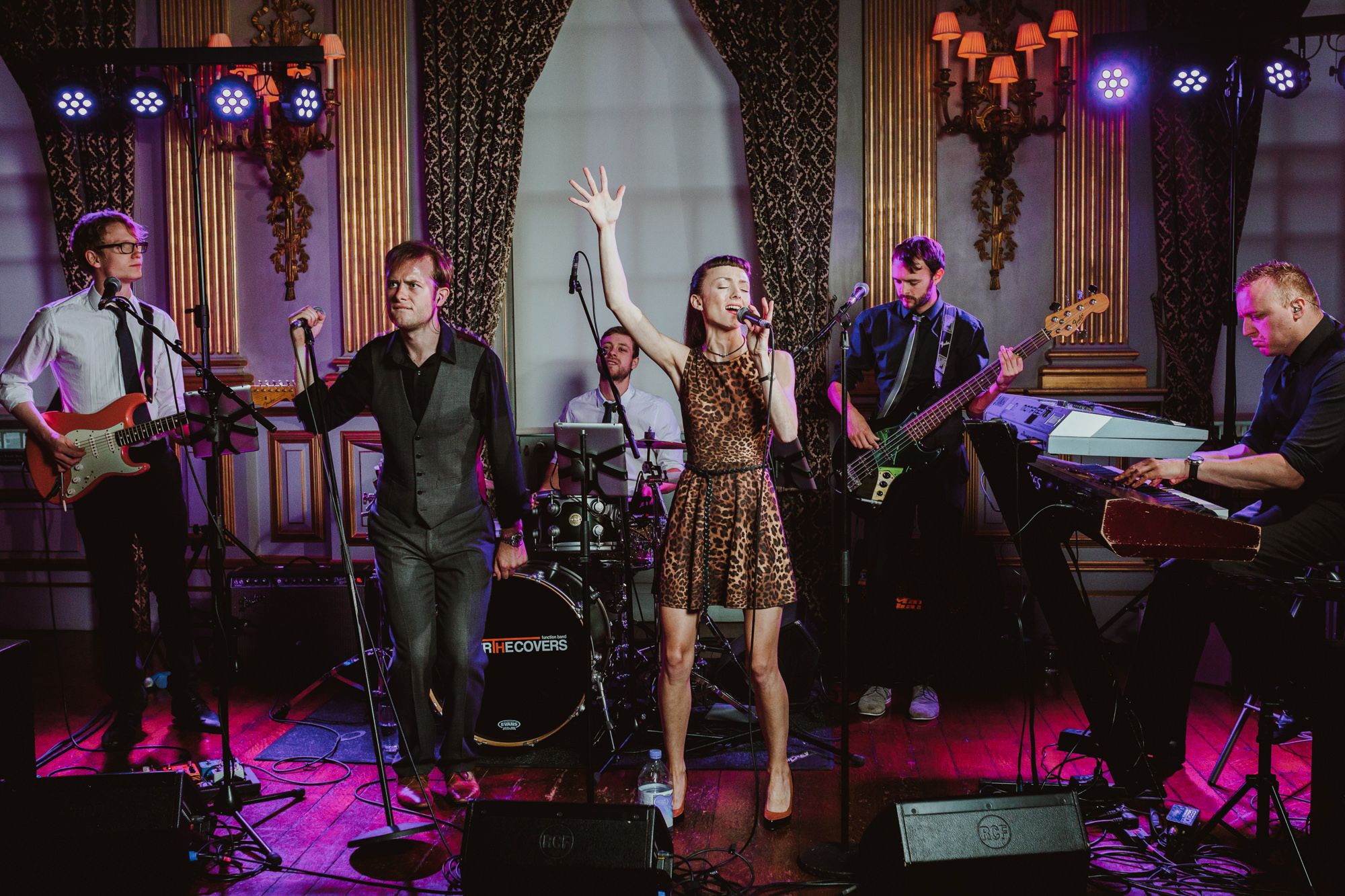 Live band performing on stage with female vocalist, guitarist, drummer, and keyboardist in elegant venue with purple lighting.