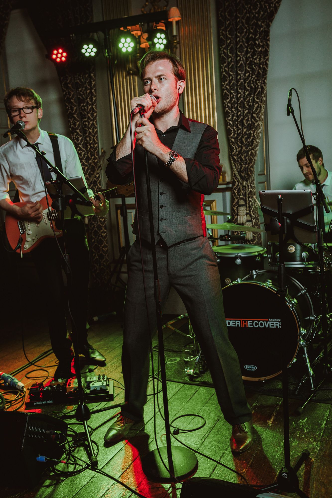 Live band performs on stage, vocalist in dark suit sings into microphone with guitarist and drummer under colorful stage lights.