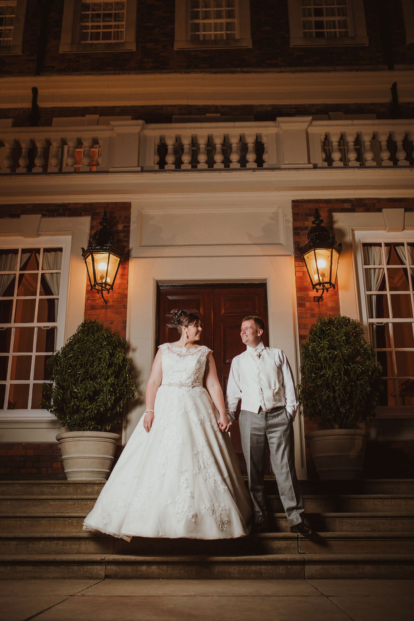 Bride and groom holding hands on steps of elegant venue at night, illuminated by warm lanterns.