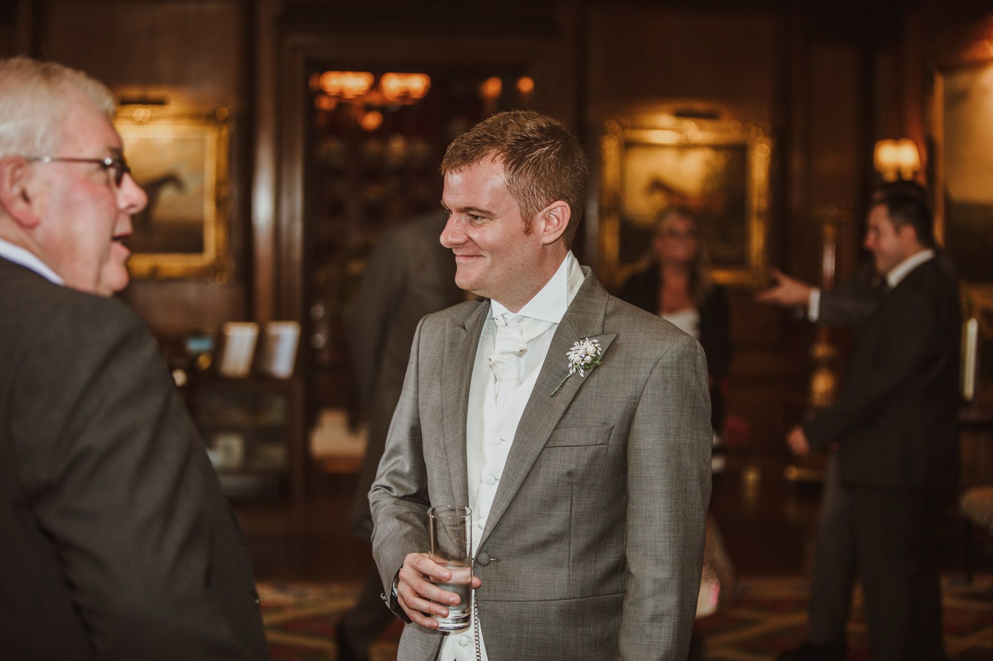 Groom in gray suit with boutonniere smiling at wedding reception in elegant wood-paneled venue.