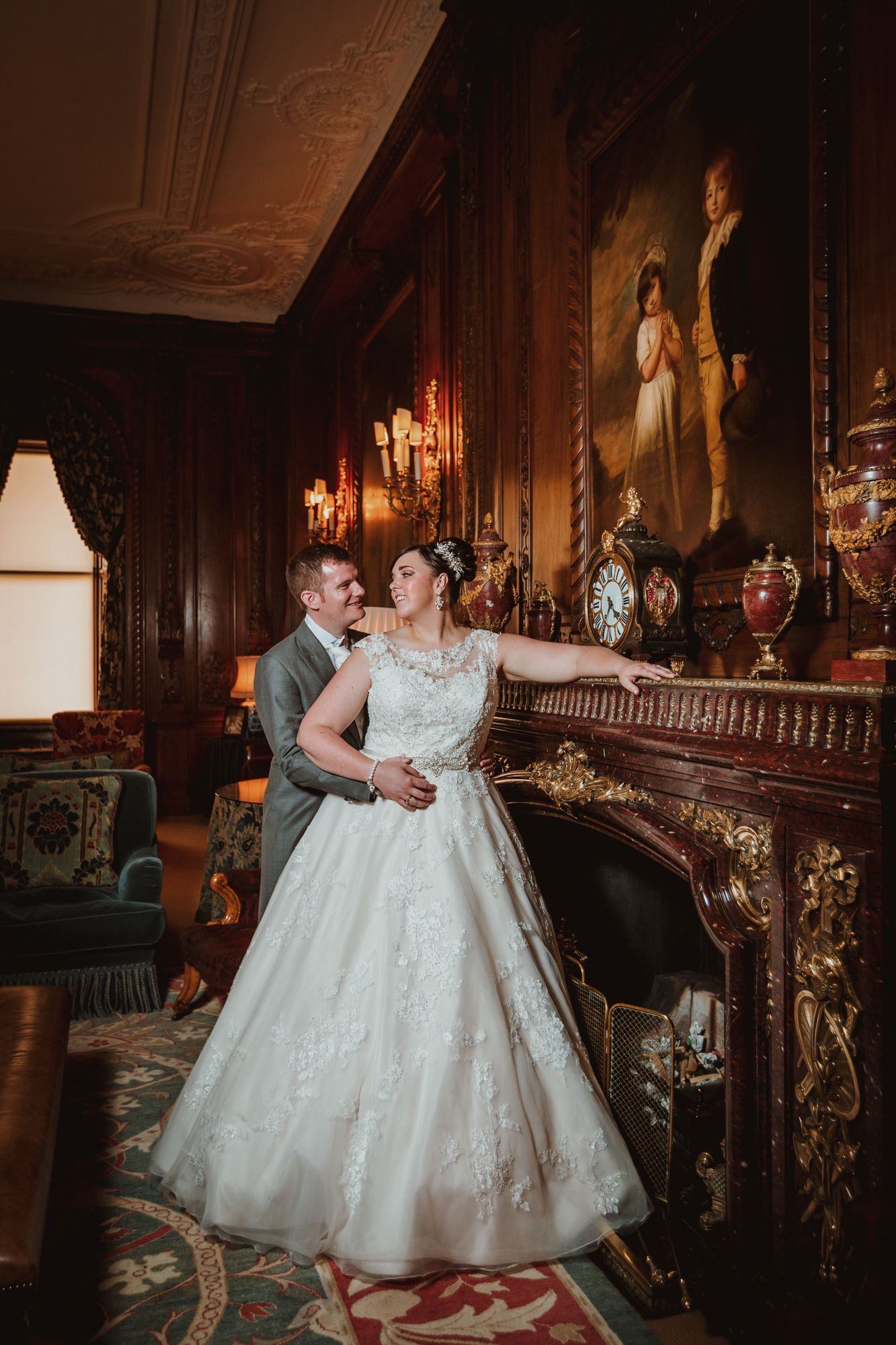 Bride in lace ball gown and groom in gray suit pose by ornate fireplace in elegant manor wedding venue.