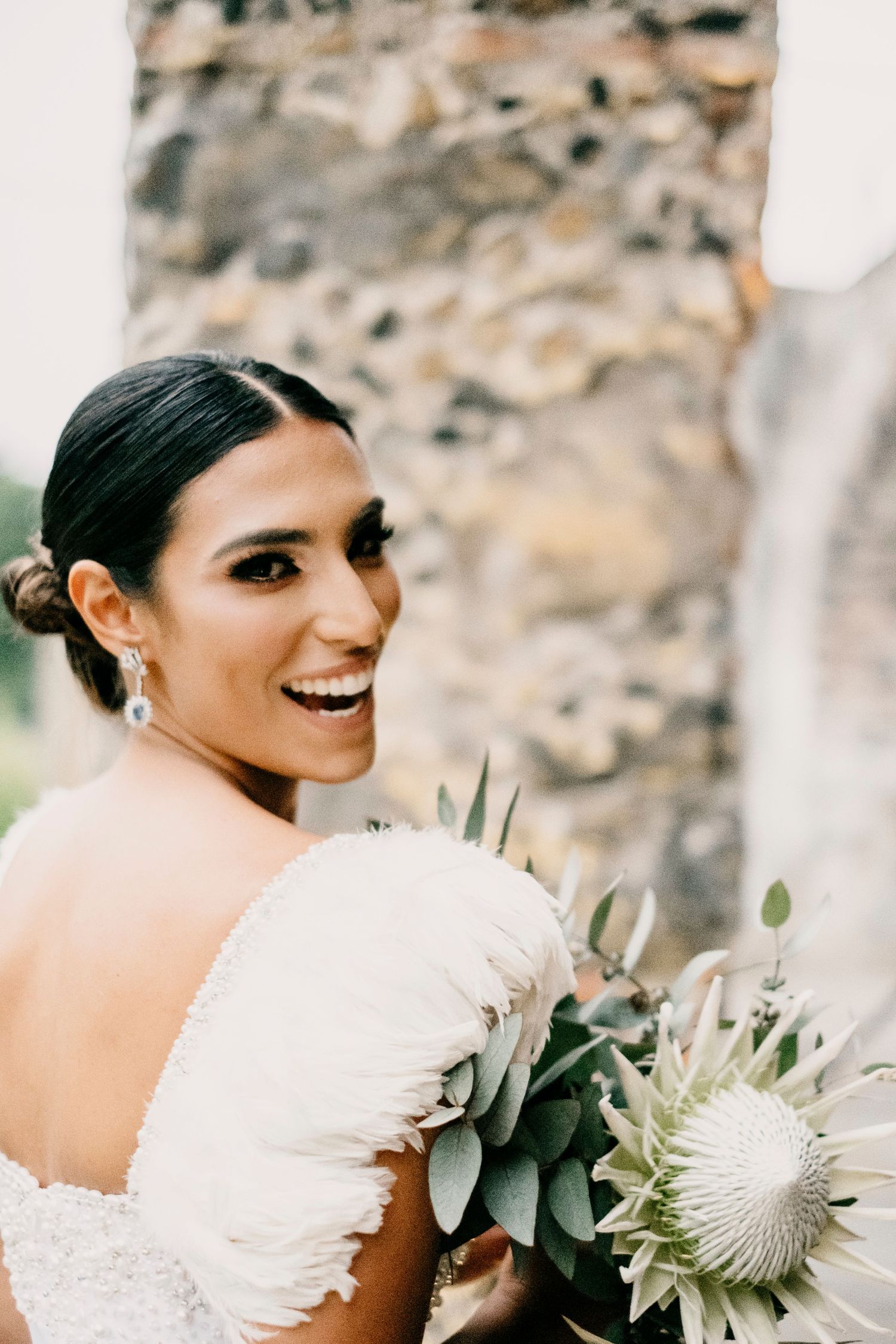 Smiling bride in white off-shoulder gown holds a white king protea bouquet with eucalyptus, posing before a stone wall.