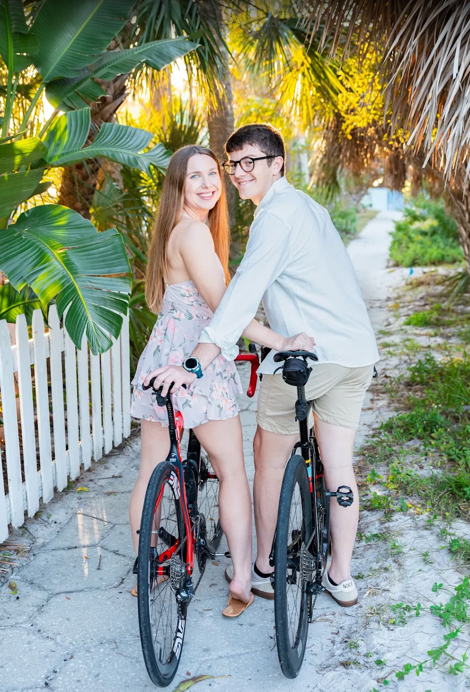 Smiling couple posing with red bicycles on a sunny tropical path lined with palm trees and a white fence.