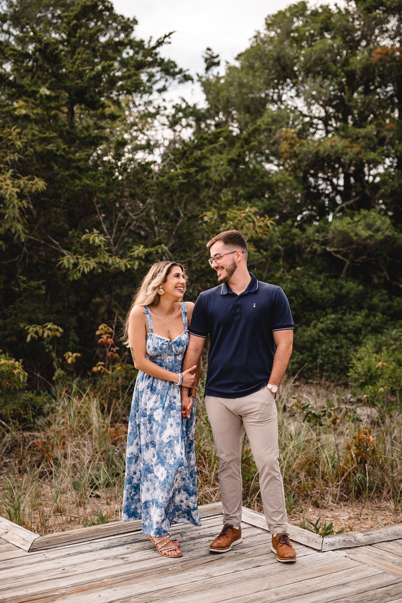 A couple smiles at each other on a wooden boardwalk surrounded by lush greenery, woman in floral dress, man in navy polo.