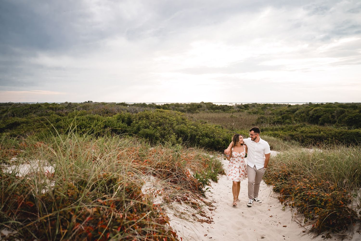 Couple walking hand in hand on a sandy beach path surrounded by lush green dunes under a cloudy sky.